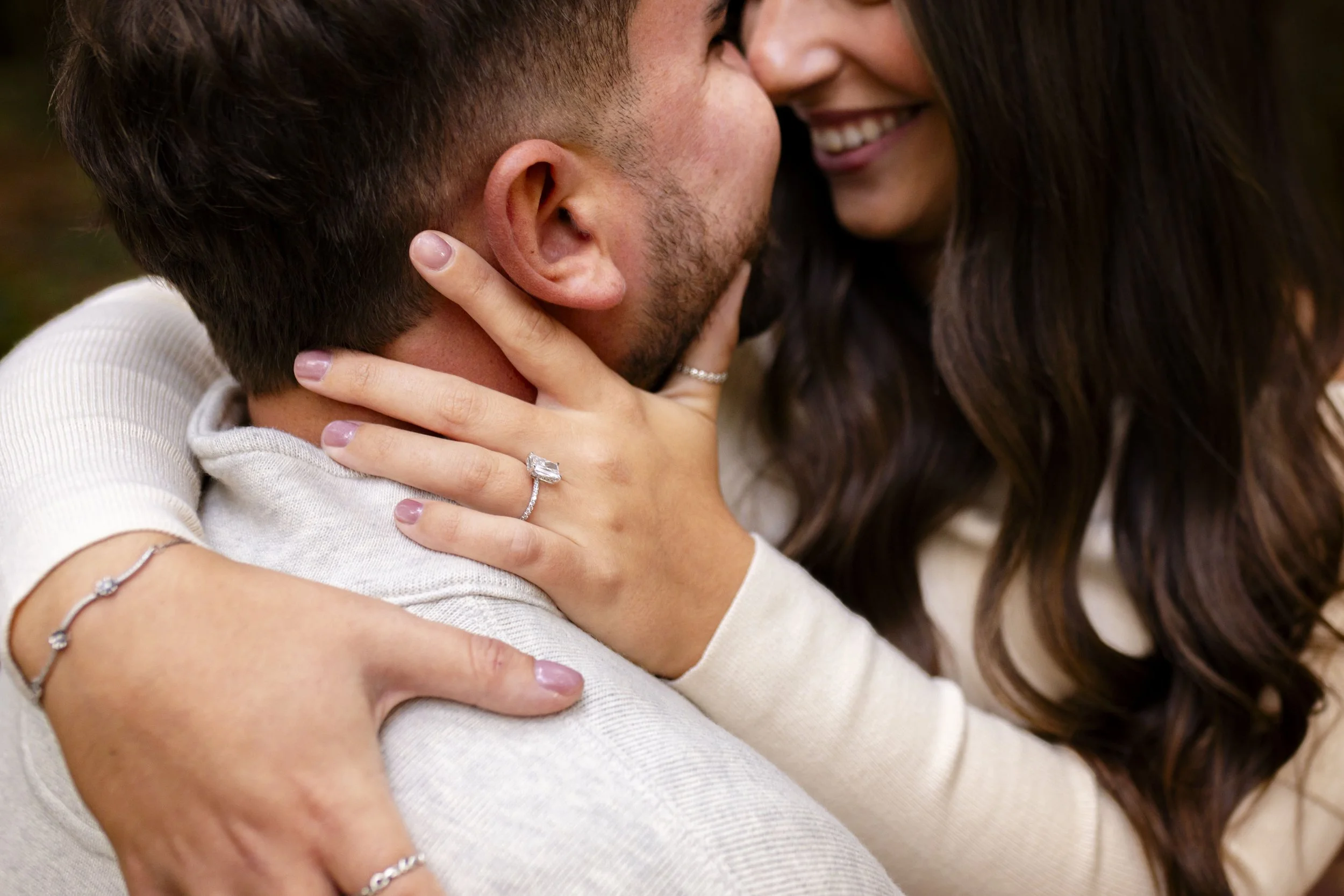 A couple close together, smiling and touching faces, with focus on their hands and rings, suggesting a romantic or engagement moment.