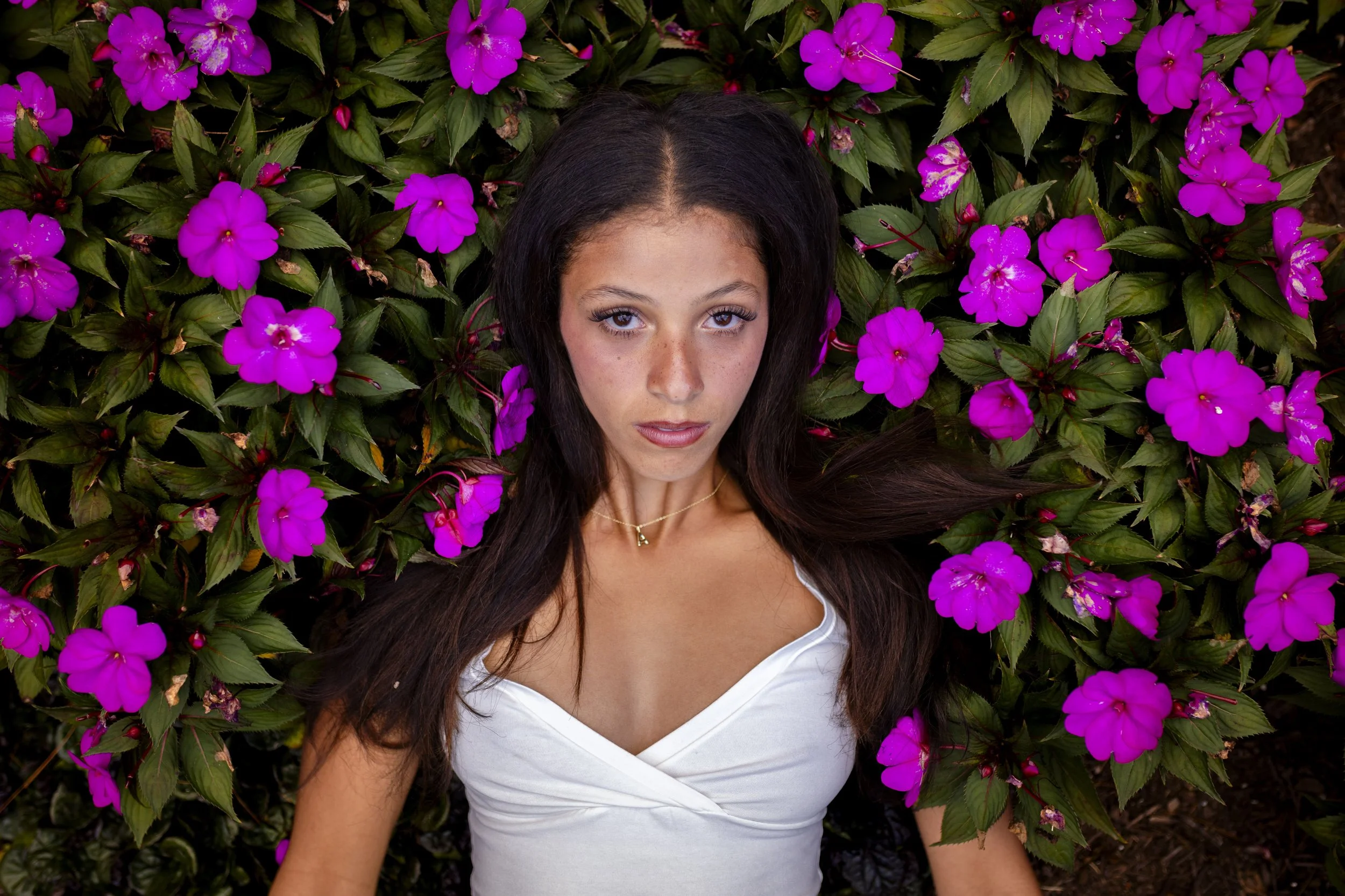 A woman with long dark hair lying on a bed of green leaves and vibrant pink flowers, wearing a white top and a gold necklace.