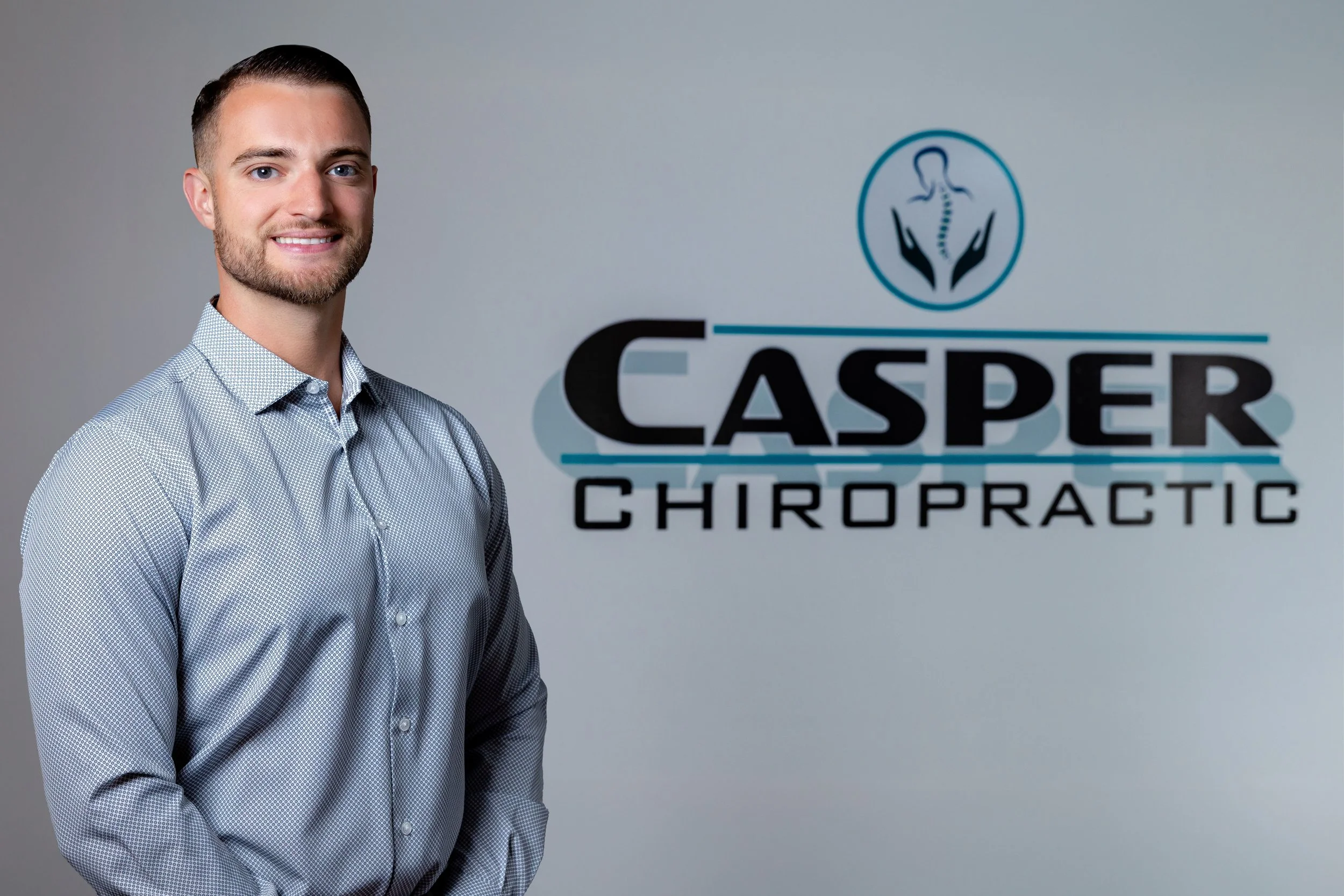 A smiling man in a light gray dress shirt standing next to a sign that reads 'Casper Chiropractic' with a logo of a spine and hands.