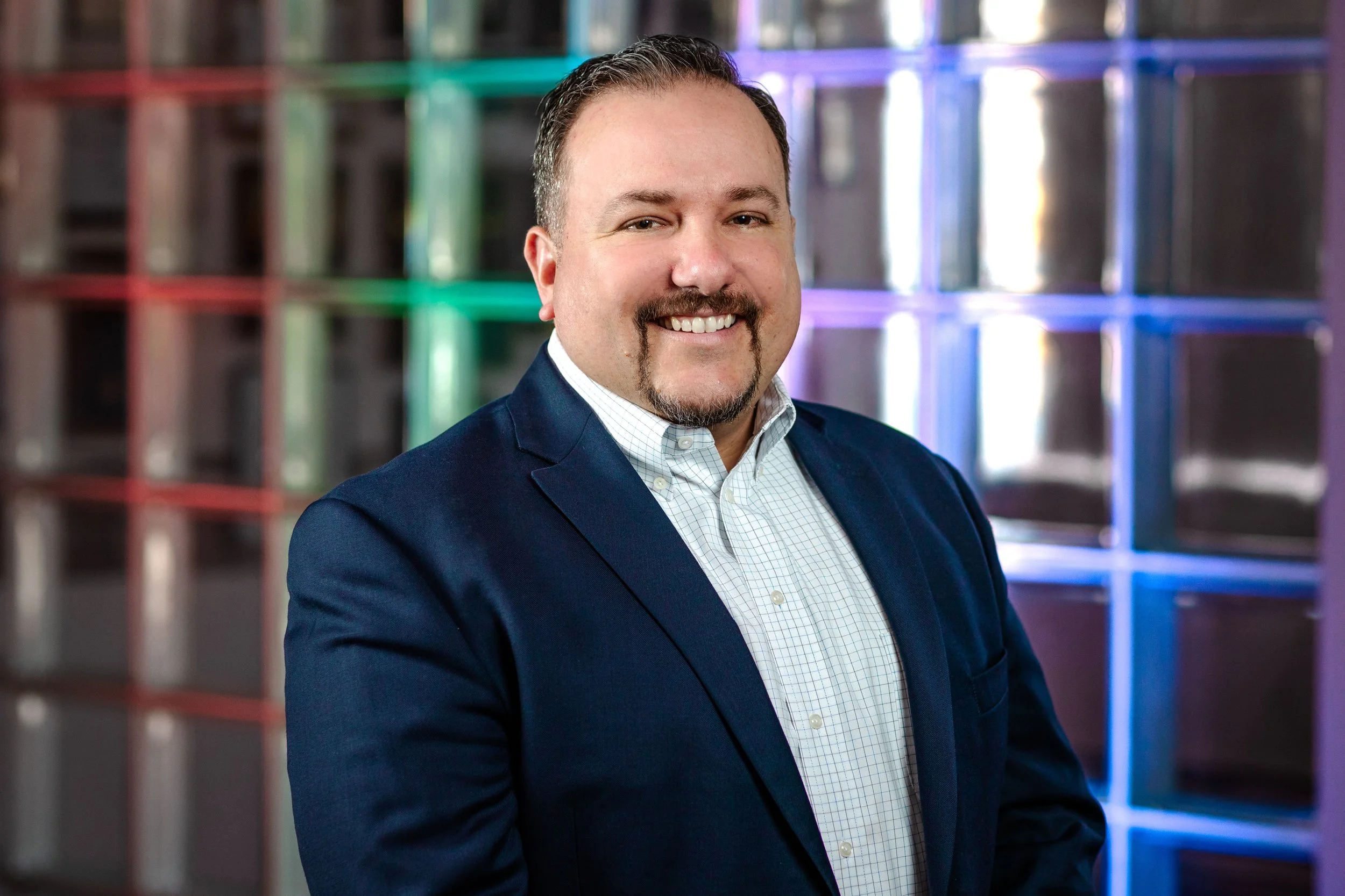 A man with a mustache and goatee wearing a navy blazer and white checkered shirt, smiling in front of a colorful illuminated geometric background.