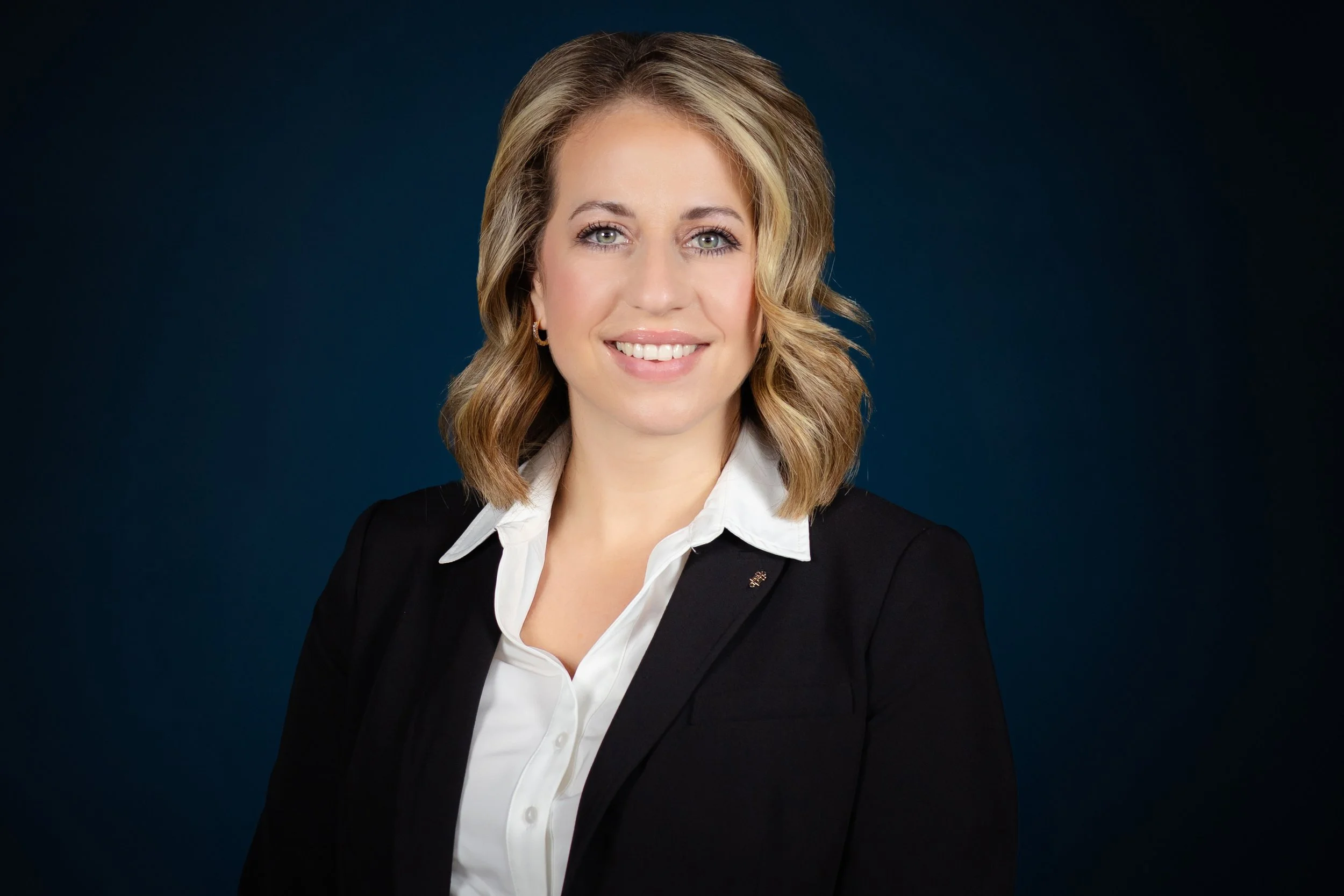 Professional woman with blonde hair smiling, wearing a white blouse and black blazer against a dark blue background.