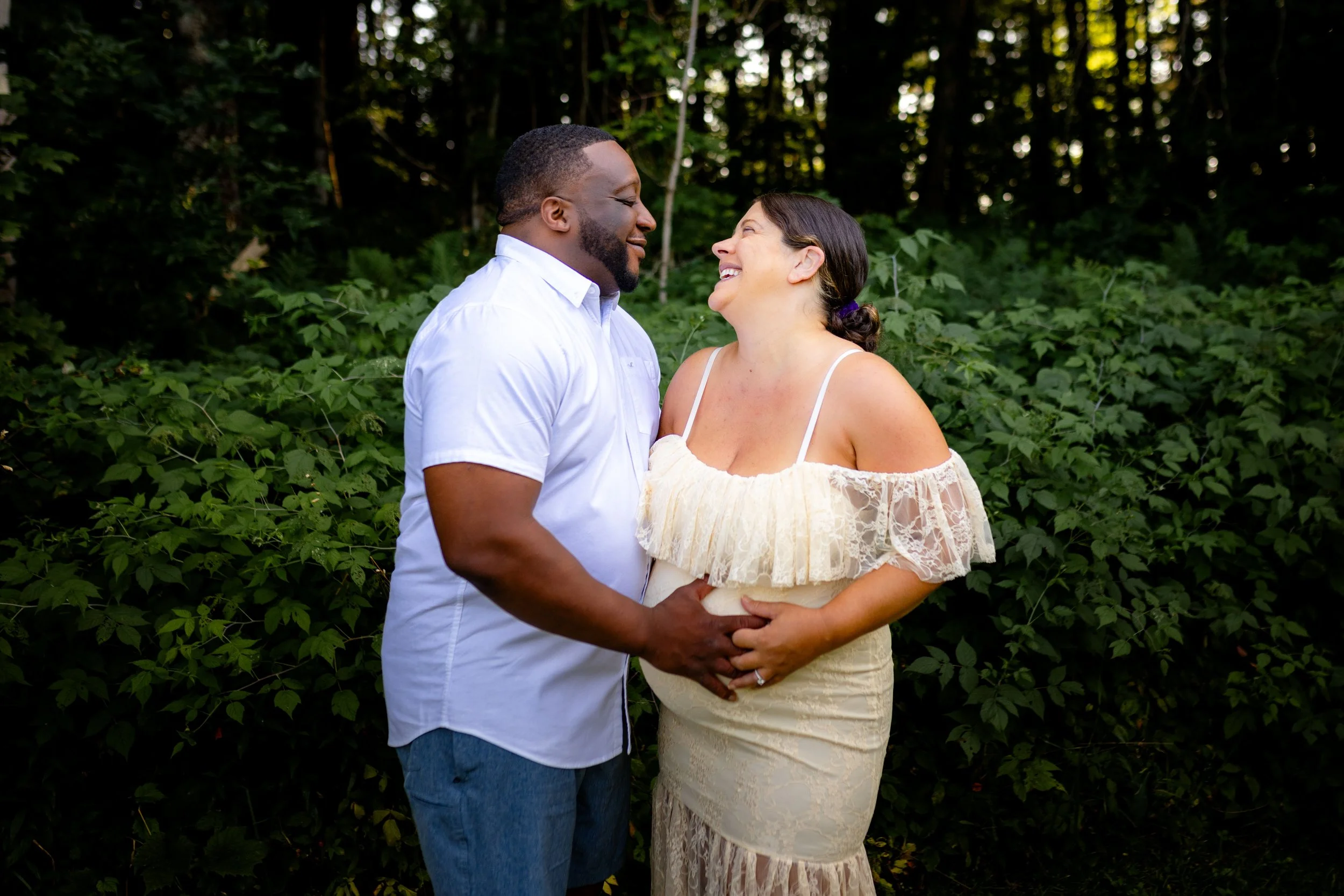 A happy couple, a man and woman, standing in front of green foliage, smiling and looking at each other. They are holding hands, with the woman’s hand resting on her pregnant belly.