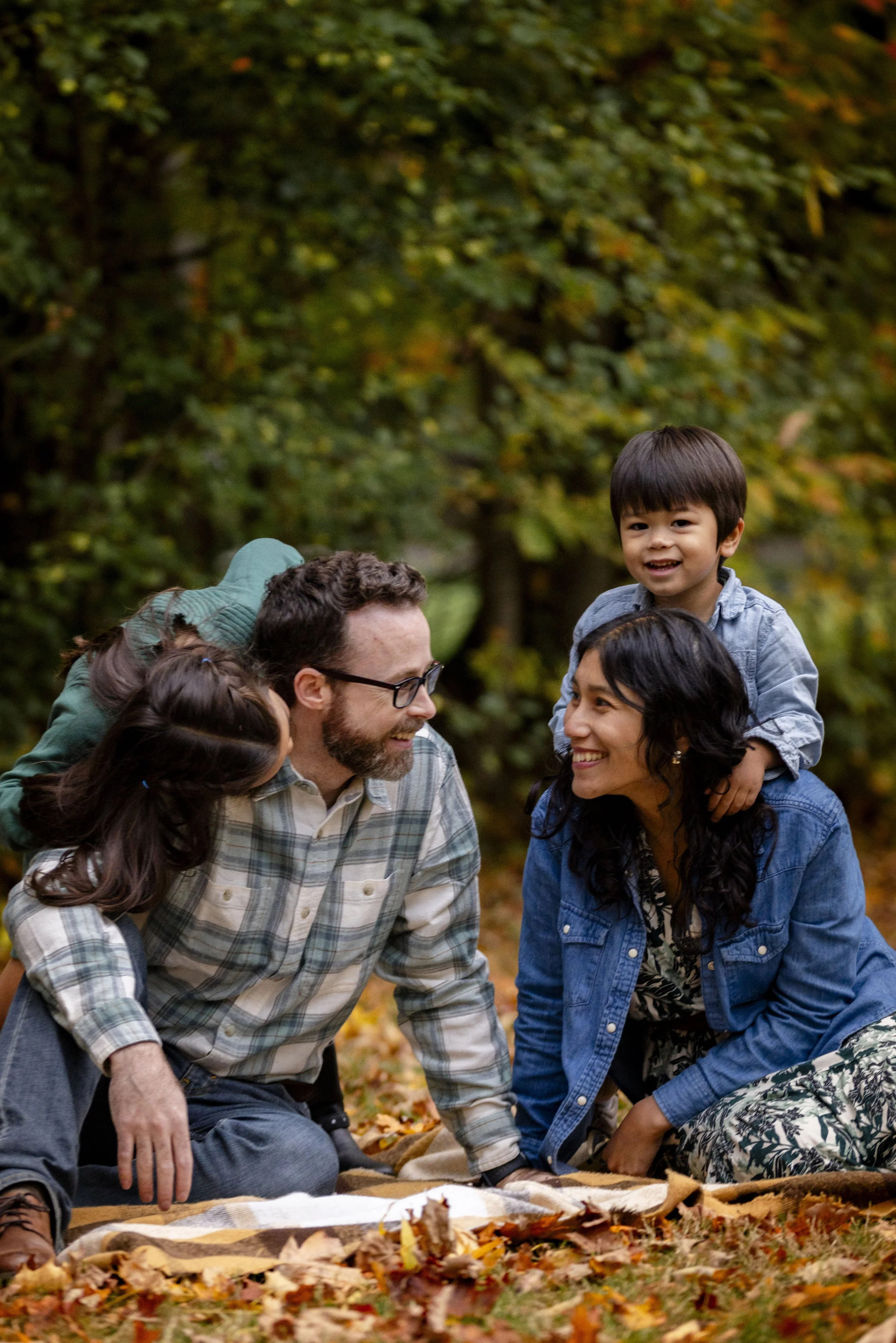 A family of four enjoying a fall day outdoors, sitting on a blanket among fallen leaves in a wooded area. The father and mother are smiling at each other while their children, a boy and a girl, are on their backs.