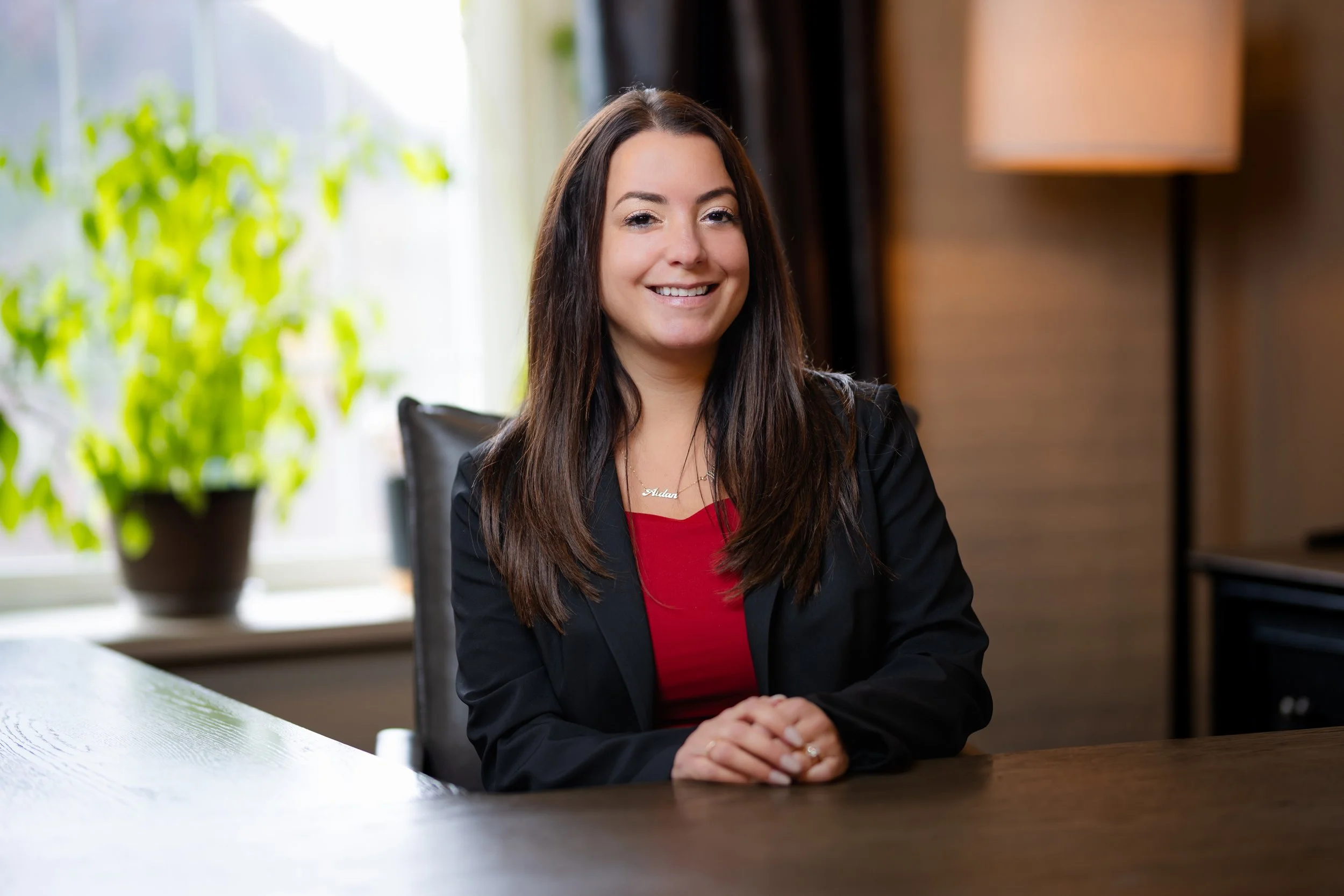 A smiling woman with long dark hair, wearing a black blazer and red top, sitting at a wooden table in a well-lit room with a potted plant and a window in the background.