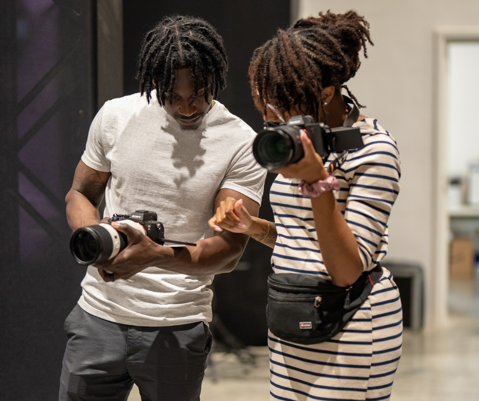 A man and a woman, both with dreadlocked hair, are looking at cameras they are holding. The woman is taking a photo with a professional camera and is wearing a striped dress and a waist pouch. The man is showing her something on his camera or phone and is dressed in a plain white t-shirt and gray pants.