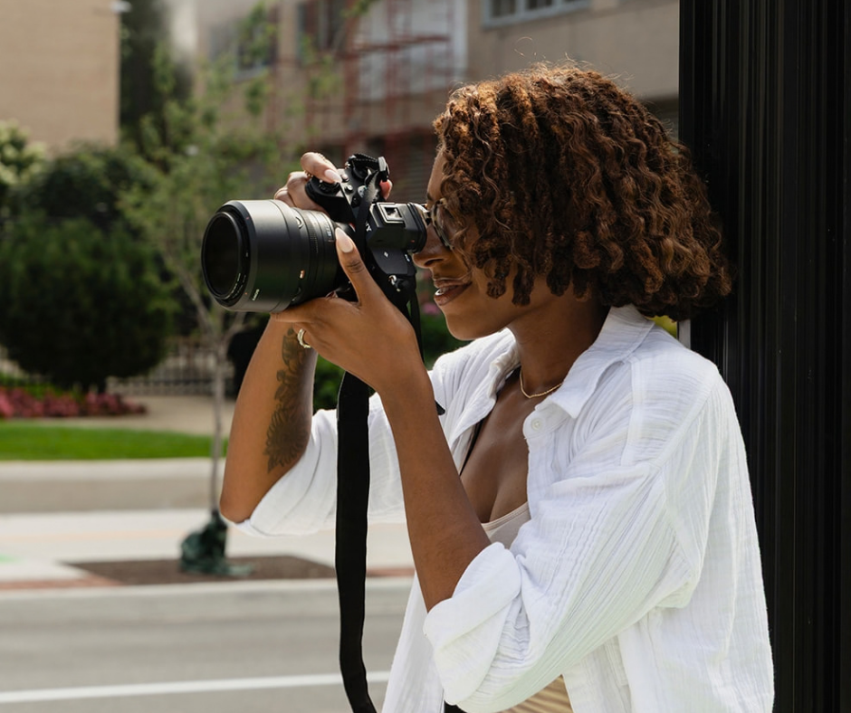 A woman with curly hair wearing glasses and a white shirt taking a photograph with a camera outdoors, next to a black fence, with trees and buildings in the background.