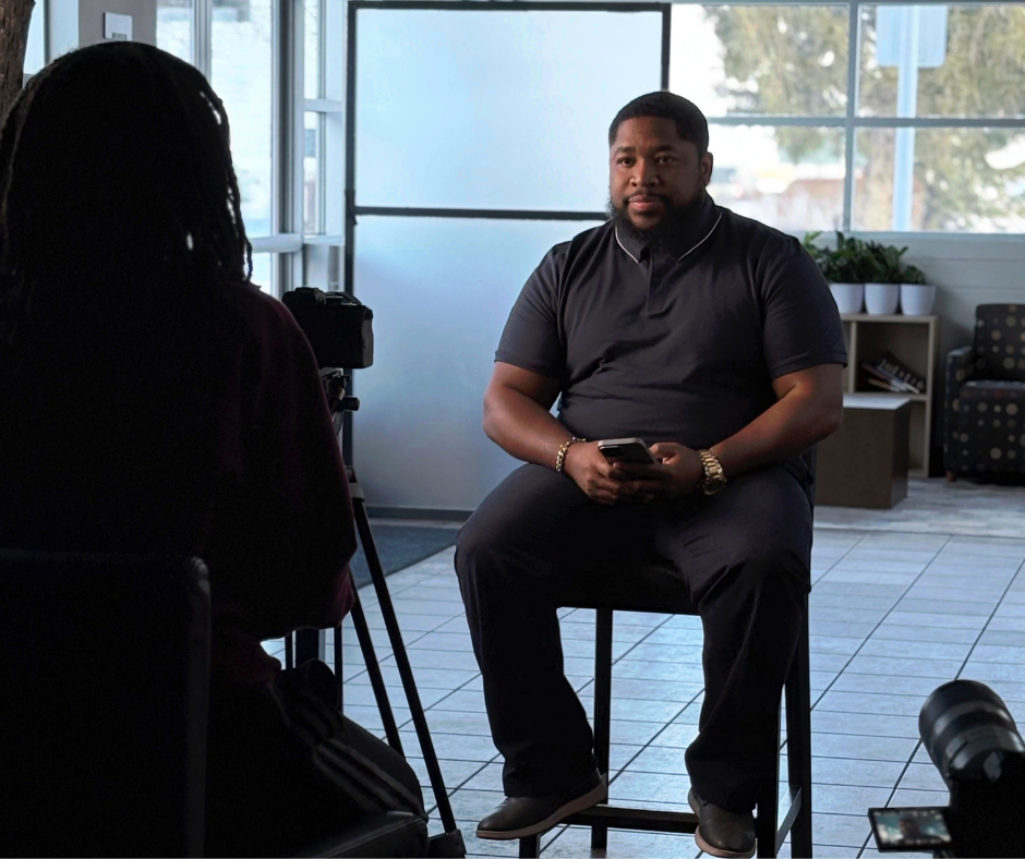 A man being interviewed in a well-lit room with large windows, sitting on a stool while holding a phone, with a camera recording him and a woman interviewer partially visible in the foreground.