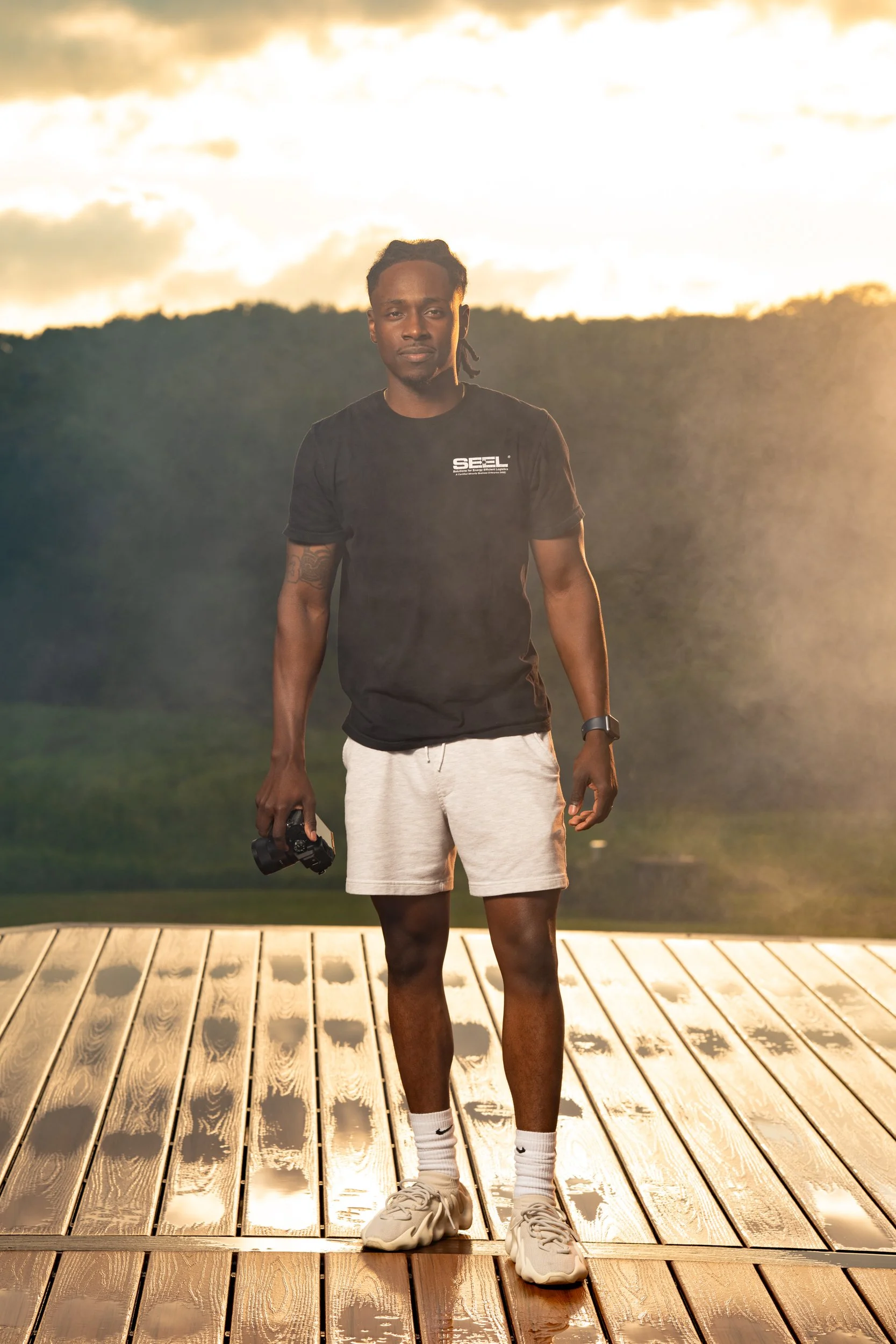 A young man standing on a wooden dock at sunset, holding a camera in his right hand, wearing a black t-shirt with a logo, beige shorts, white socks, and sneakers.