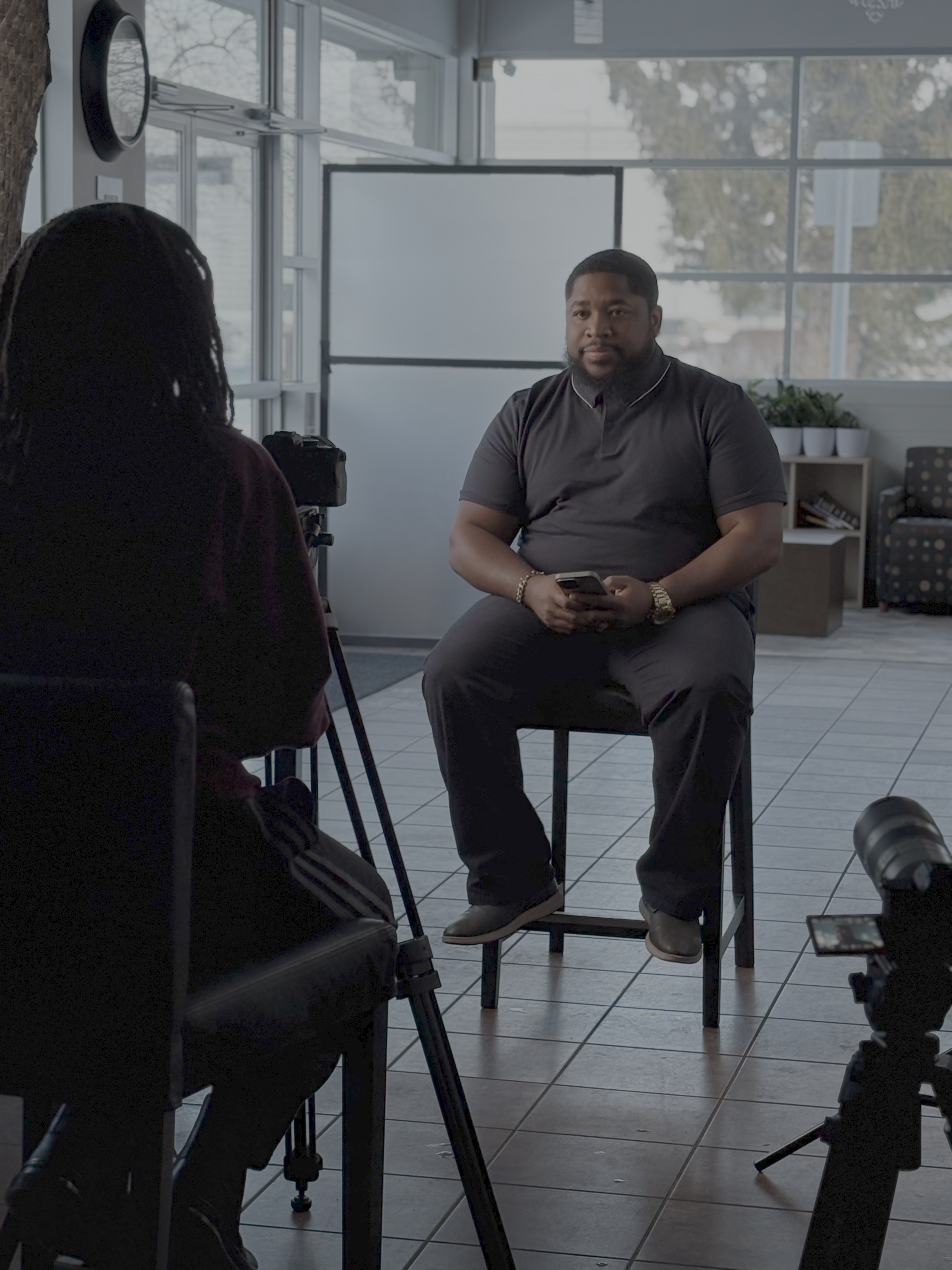 A man is seated in a chair holding a smartphone, being interviewed by a person with dreadlocks who is camera-shooting him. The setting is indoors with large windows behind, revealing trees outside. There is a whiteboard and some potted plants in the background.
