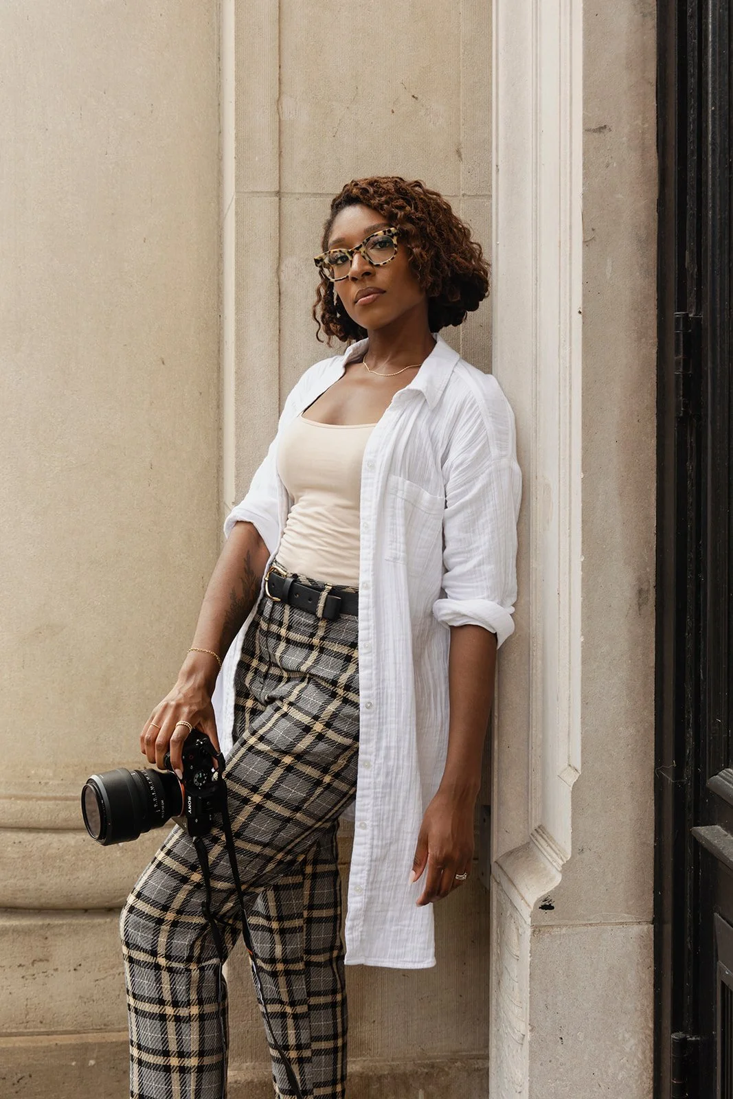 A woman with curly hair wearing glasses, a white shirt over a beige top, plaid pants with a black belt, holding a camera, standing against a beige wall outside a building.