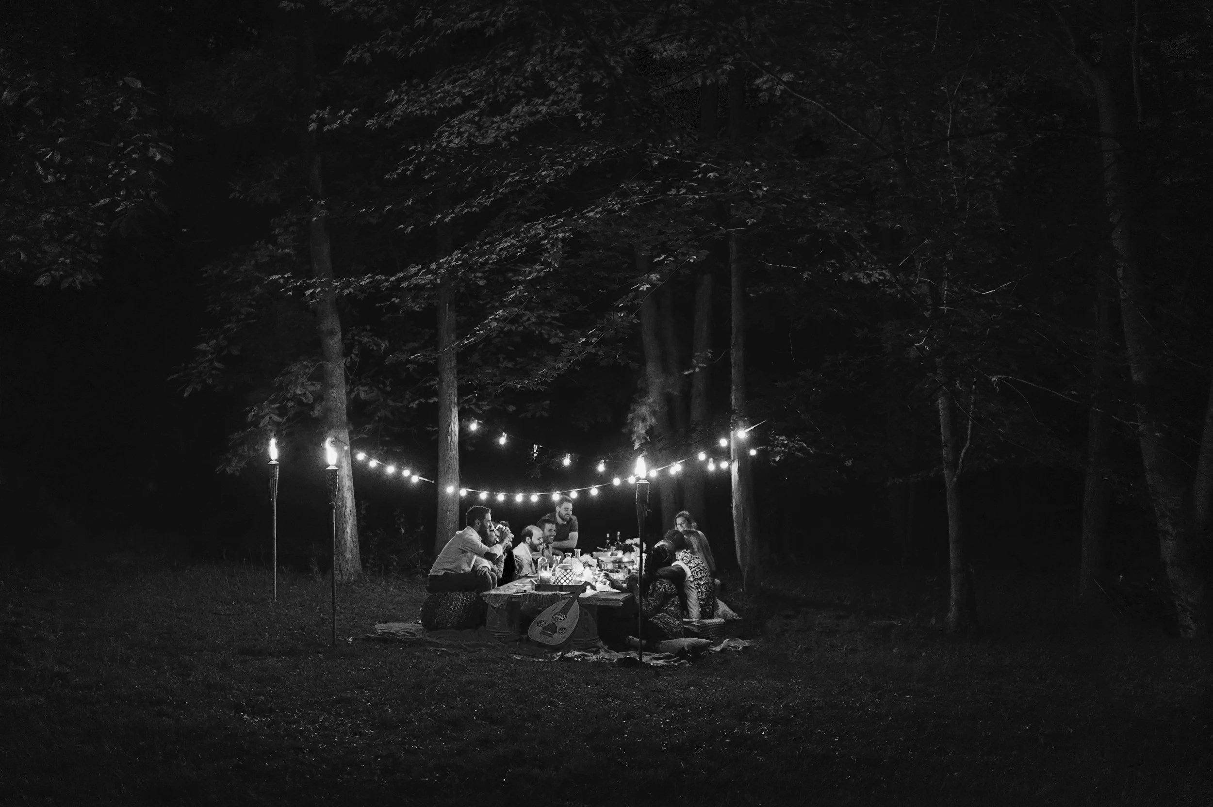A group of people gathered around a table outdoors at night, under string lights and torches, in a wooded area, having a meal or celebration.
