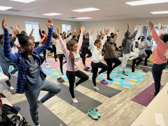 Group of people participating in a yoga class in a bright room, standing on yoga mats with arms raised in a tree pose.