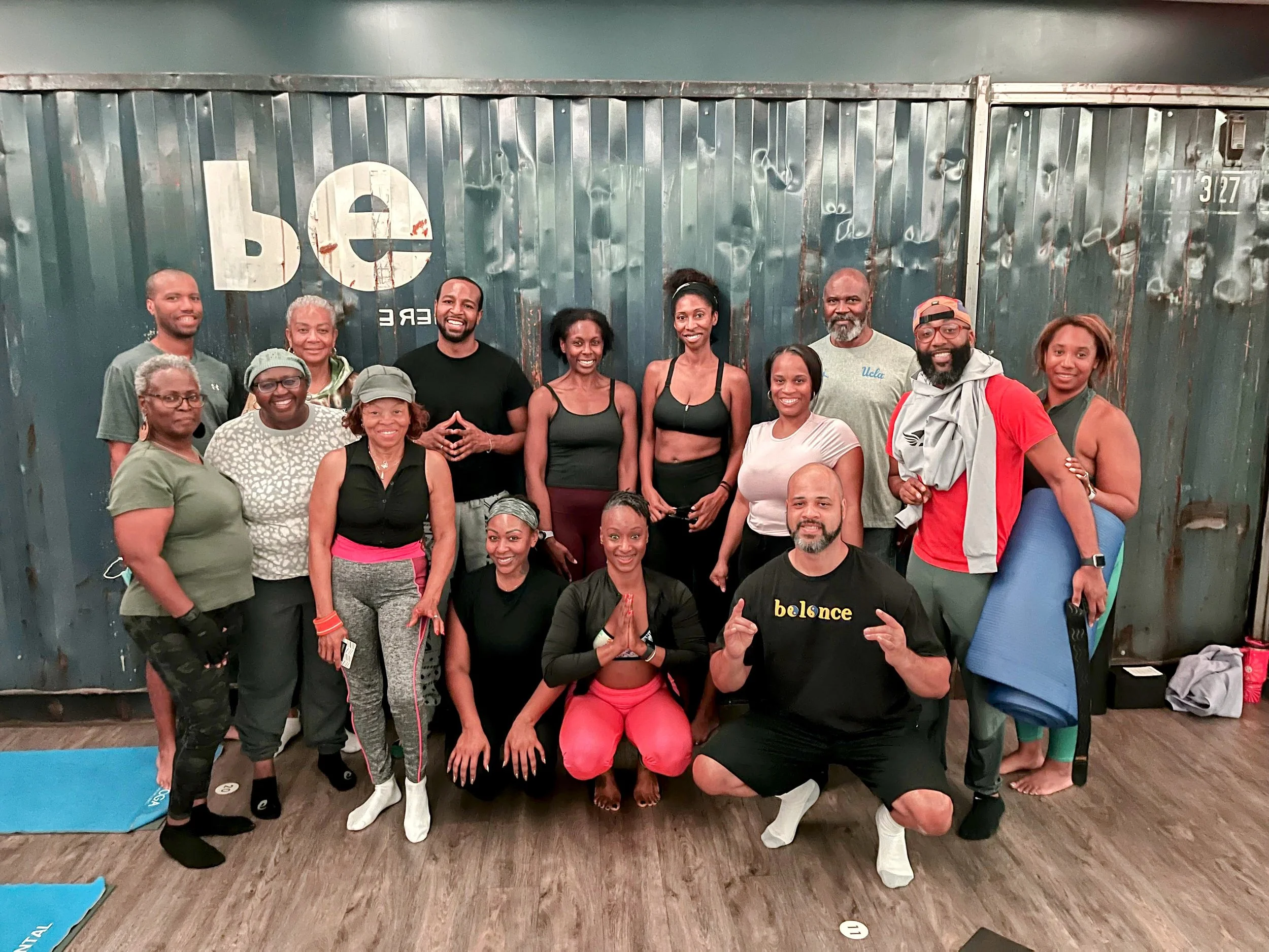 Group of people in fitness attire smiling indoors in front of a metallic wall with the 'be' logo, some holding yoga mats.