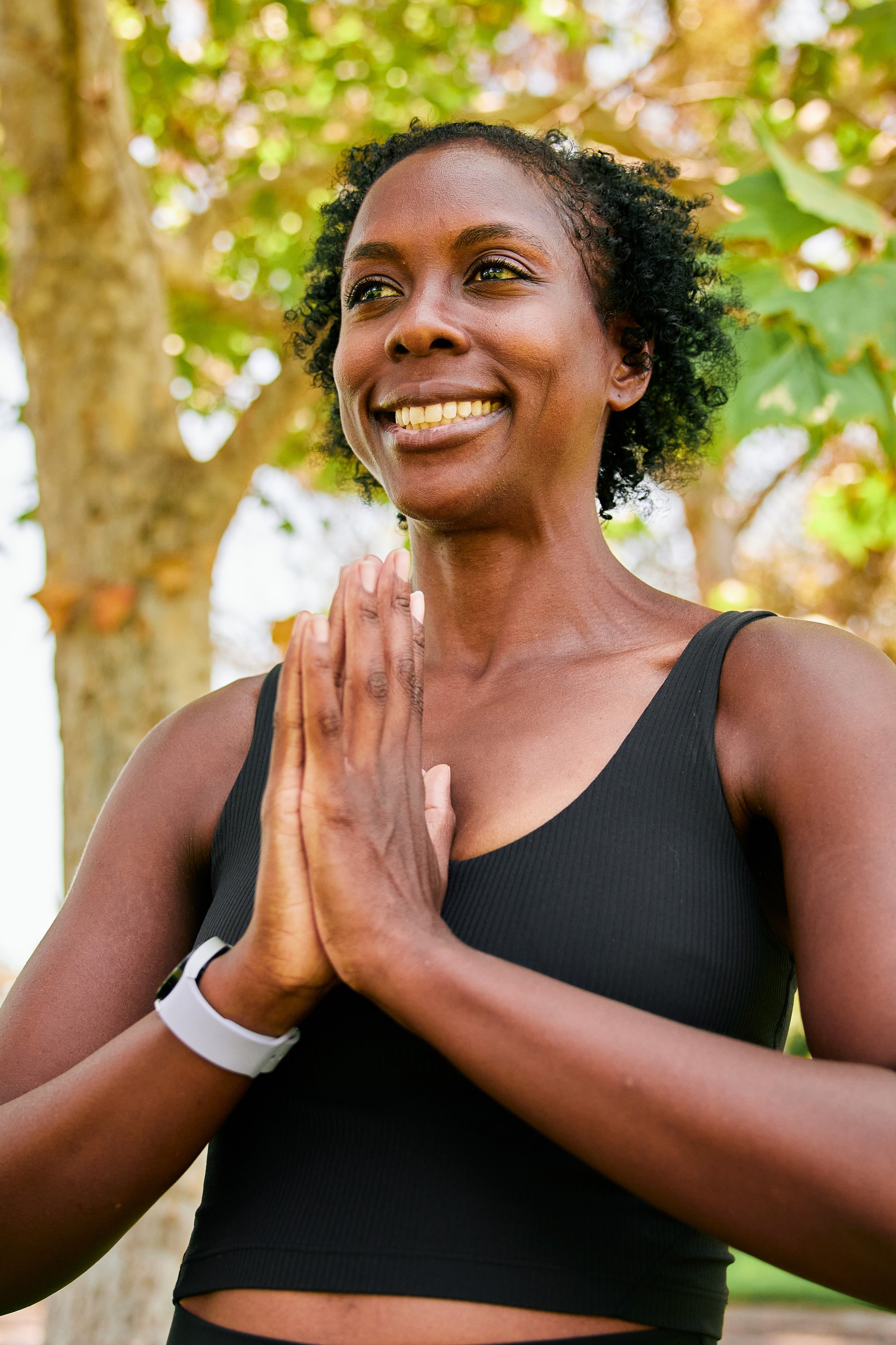 A woman with short curly hair smiling and holding her hands together in a prayer position outdoors near a tree.