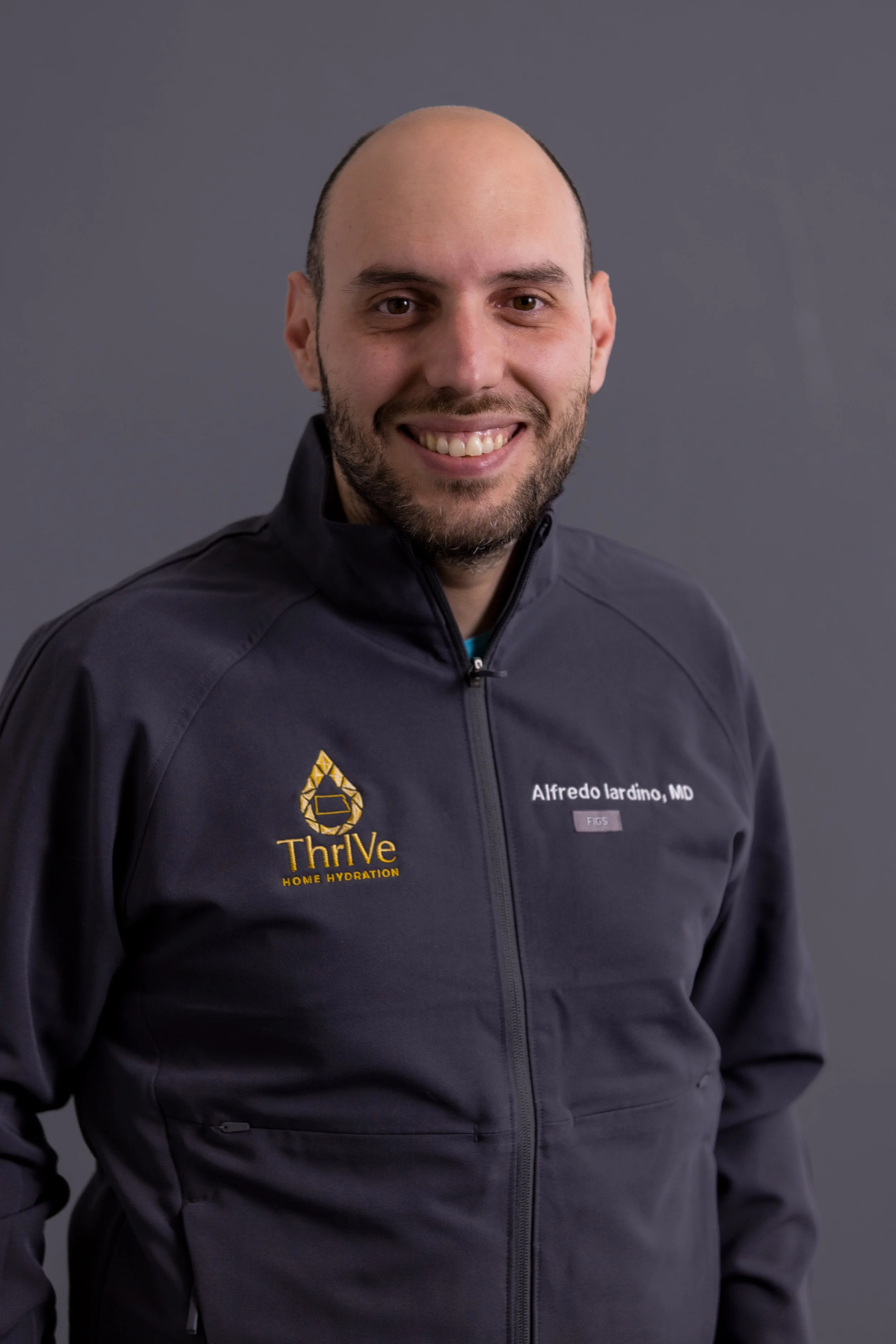 A smiling man with a beard and light skin, wearing a black jacket with a logo and a name tag that says Alfredo Iardino, MD, standing against a dark gray background.