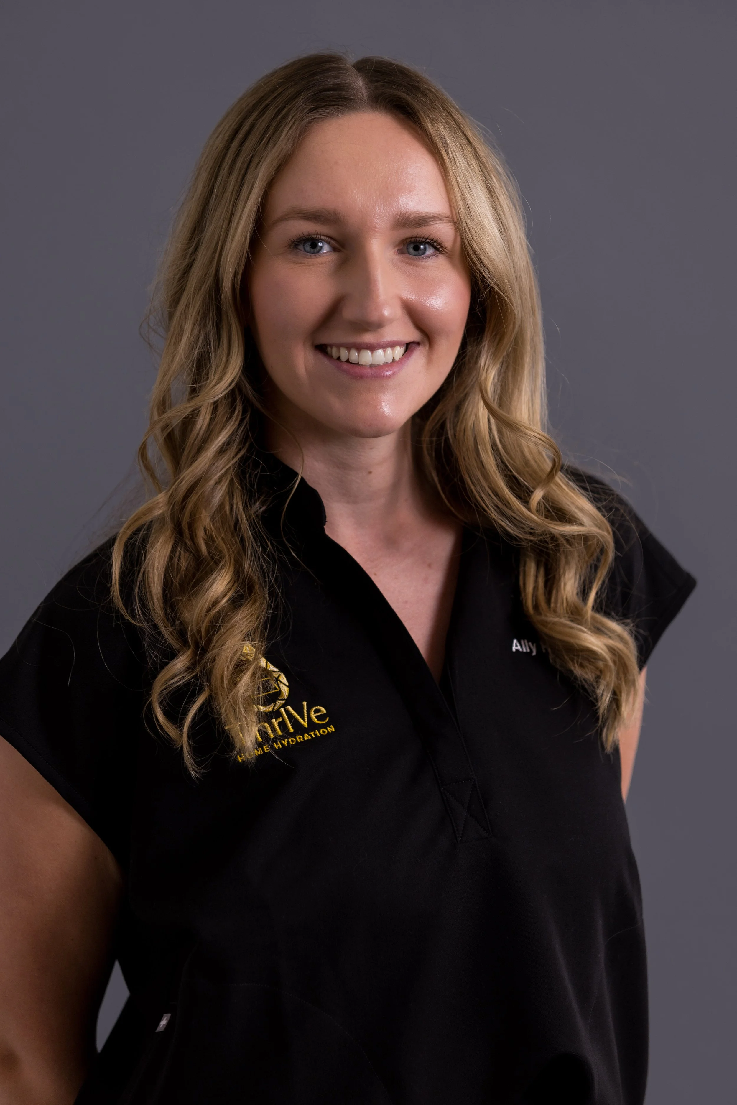 A woman with long blonde hair smiling at the camera, wearing a black shirt with gold embroidery that says 'drive' and 'home hydration' on the chest.