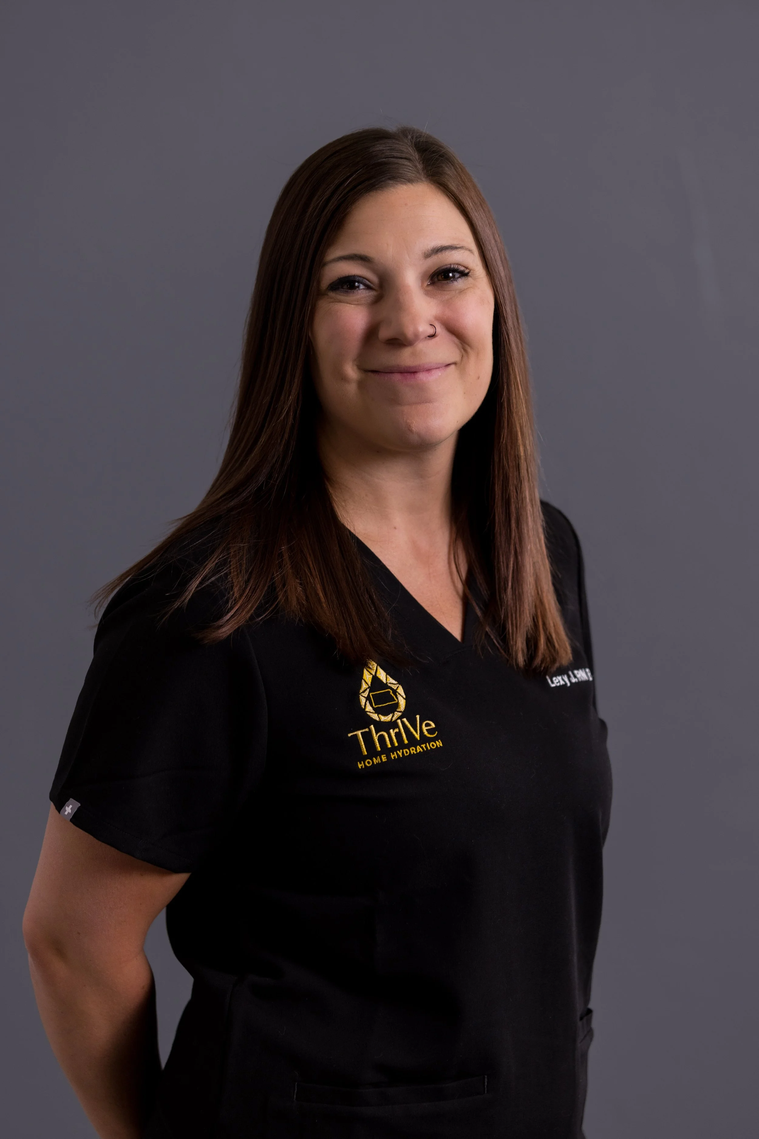 A woman with long brown hair, smiling, wearing a black Thrive Home Hydration uniform against a plain gray background.