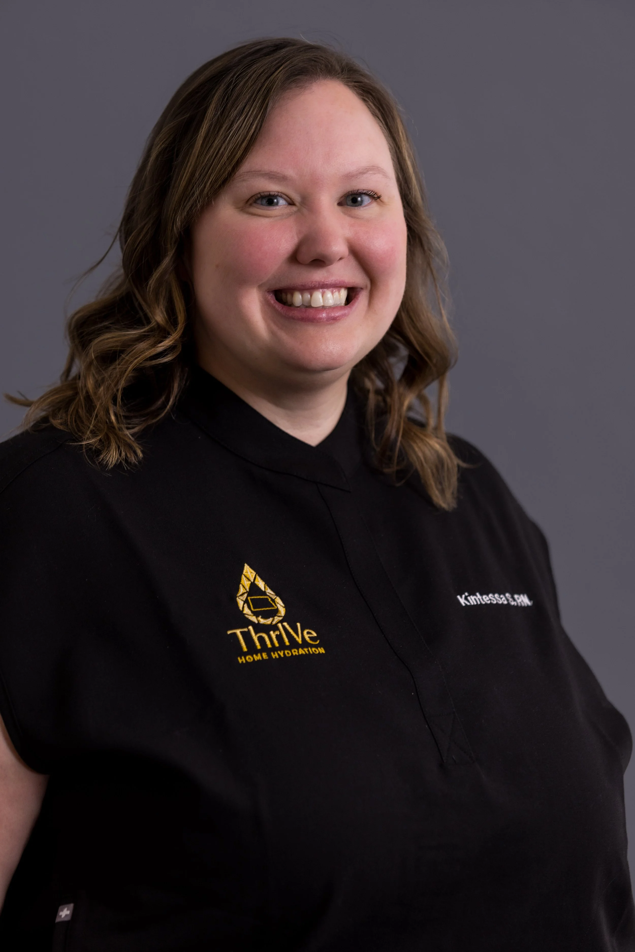 Portrait of a woman with shoulder-length wavy brown hair, wearing a black shirt with a yellow logo and the words 'Thrive Home Hydration' and her name 'Kimtessa S. PAM' embroidered on it, against a gray background.