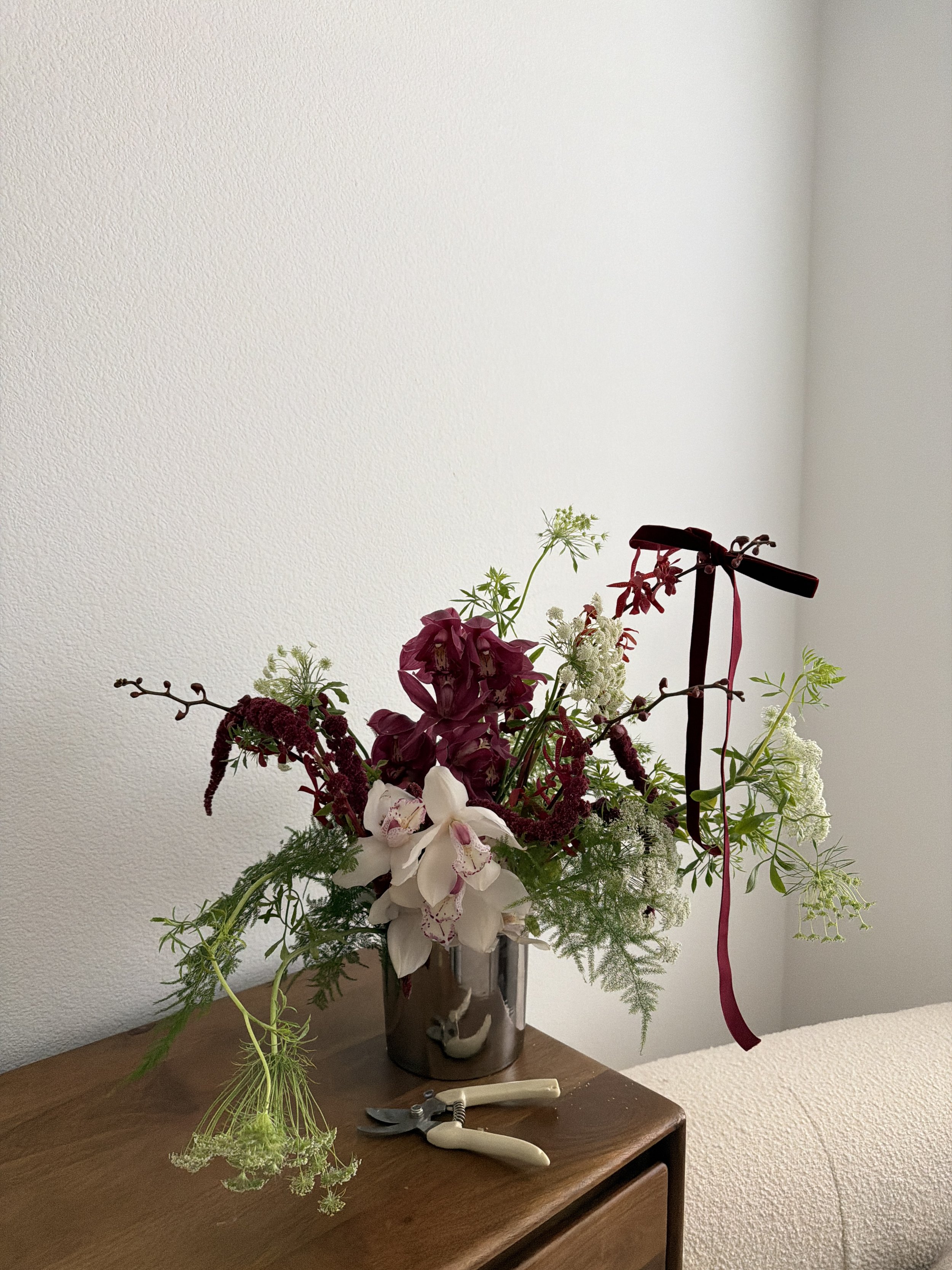 A floral arrangement in a copper-colored vase with white, pink, and dark red flowers, decorated with a dark ribbon, on a wooden surface with pruning shears nearby.