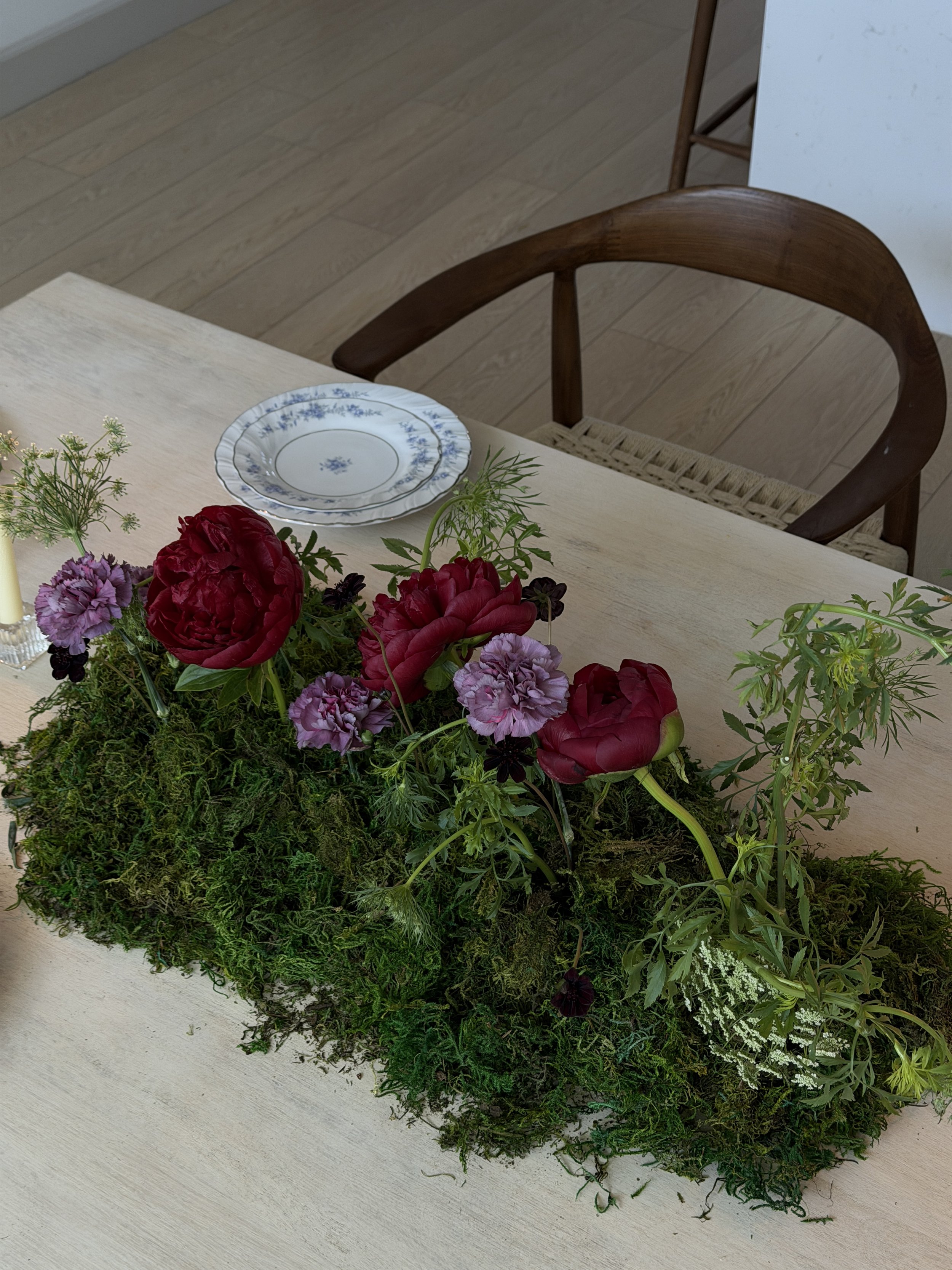 A floral arrangement of pink and purple flowers on a mossy green base on a wooden table, with a blue-and-white patterned plate and a chair in the background.