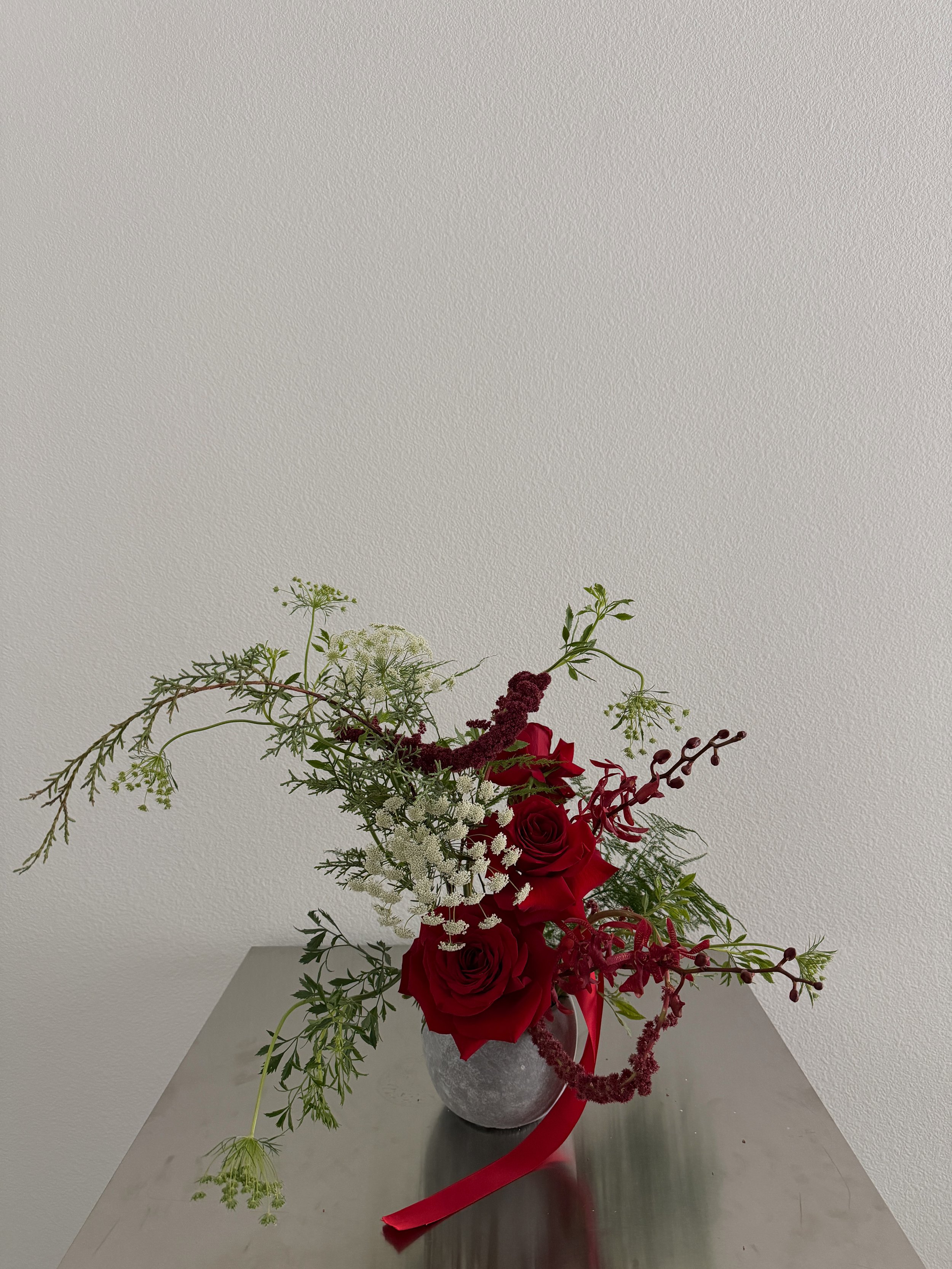 A floral arrangement with red roses, white flowers, and green foliage in a round, textured gray vase on a metallic surface.