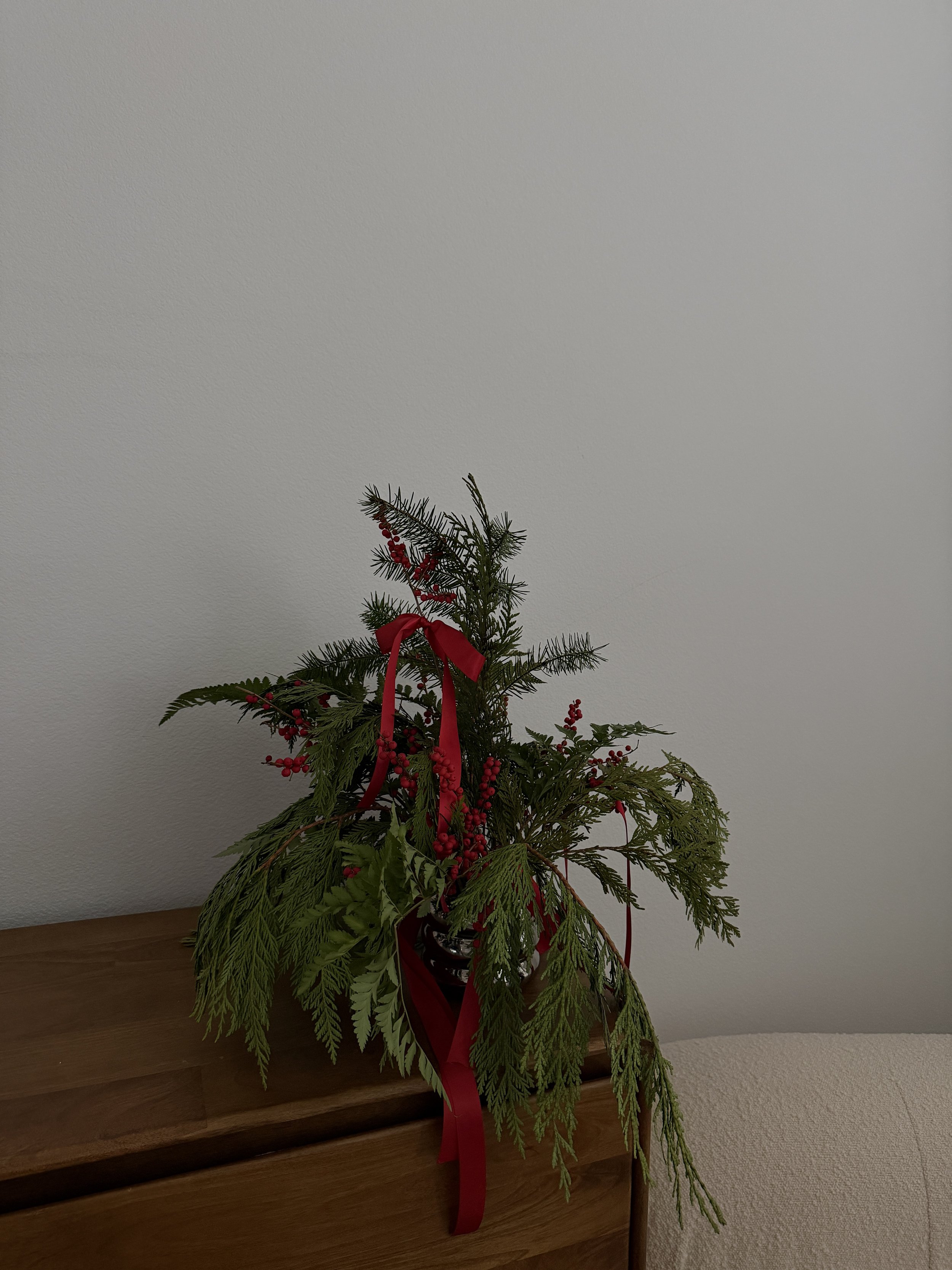 A festive holiday floral arrangement with greenery and red berries, adorned with a red ribbon, placed on a wooden surface against a plain wall.