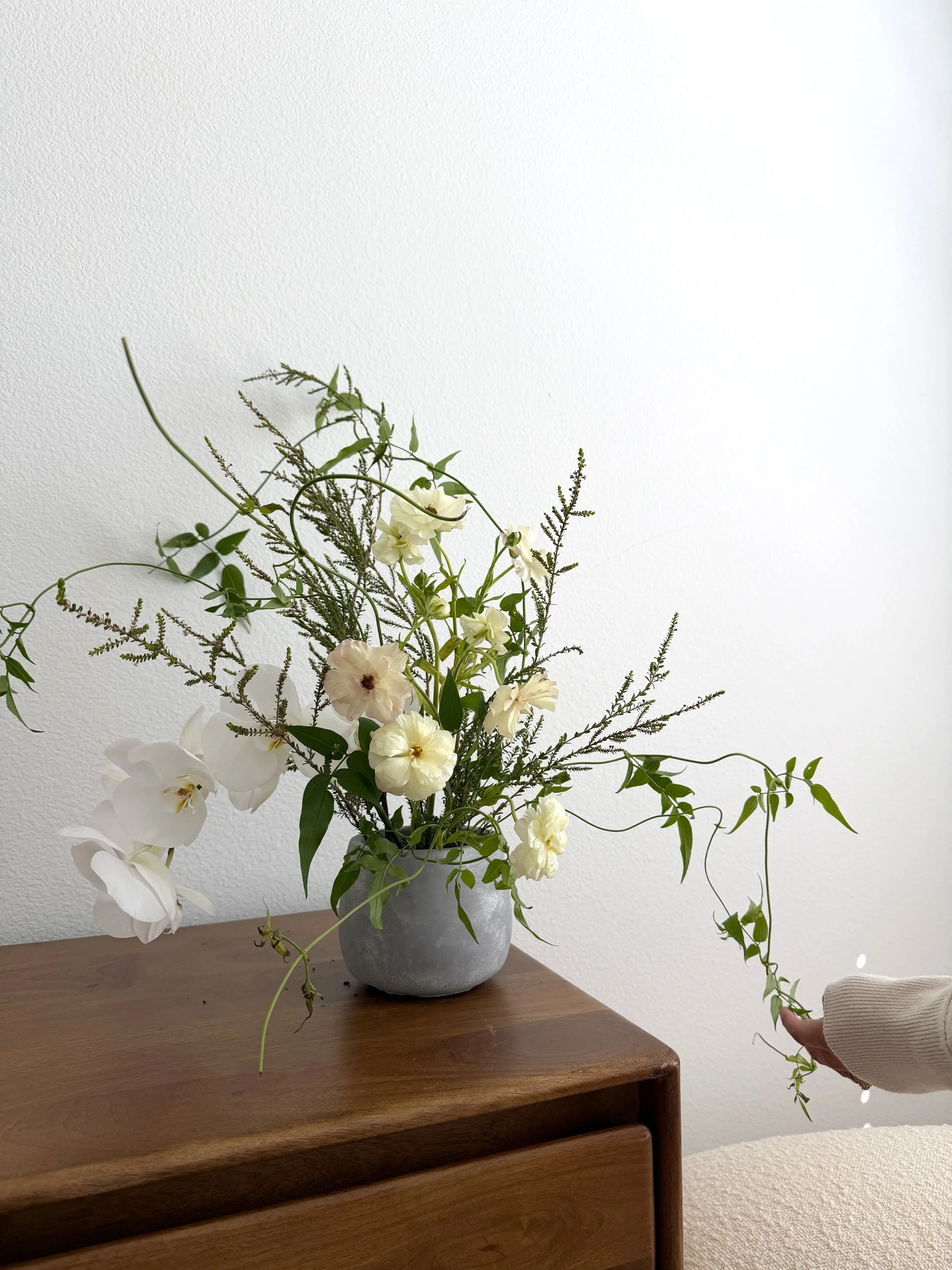 A white flower arrangement in a gray vase on a wooden table, with a person's hand touching a vine near it against a plain white wall.