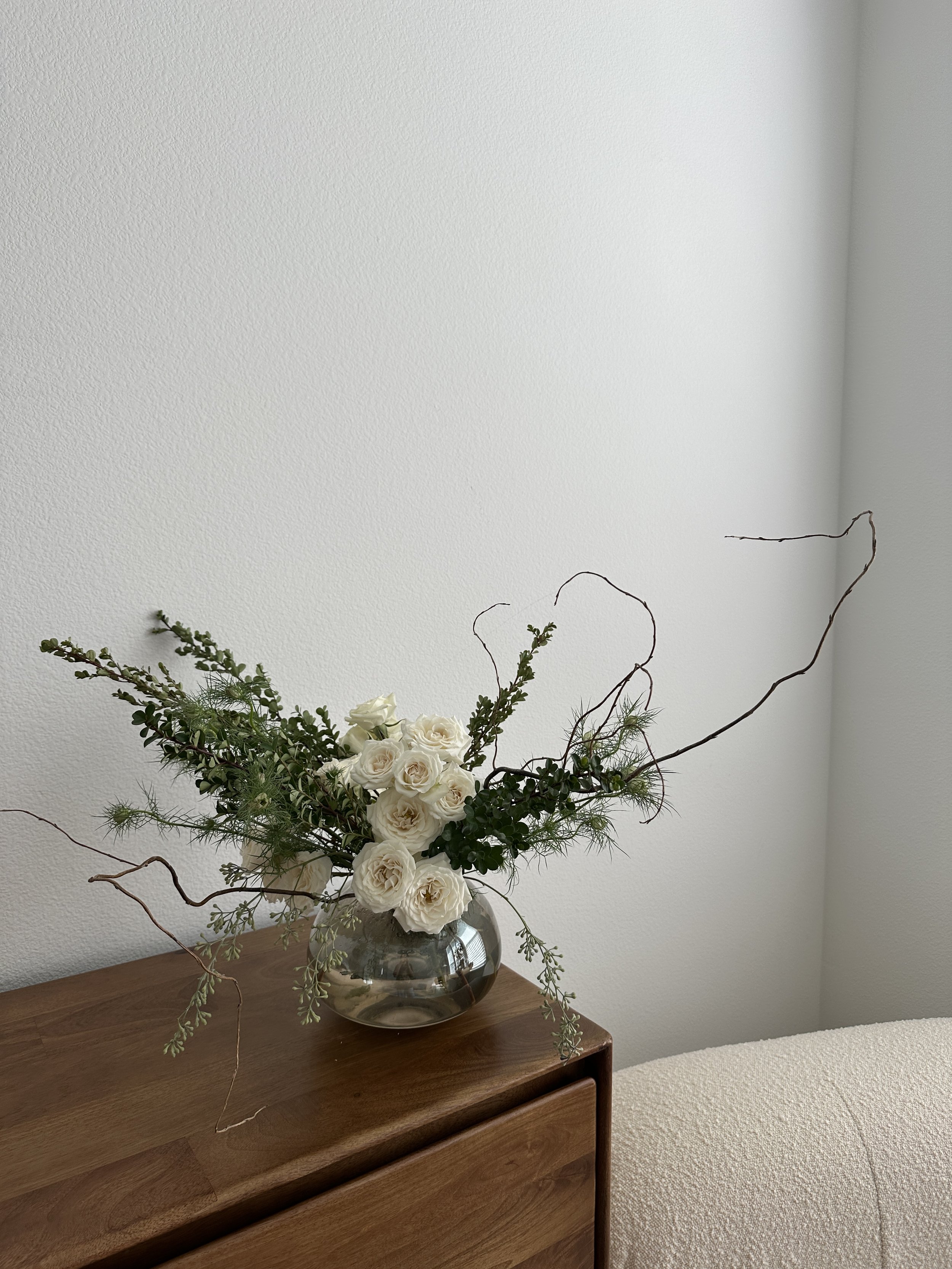 A floral arrangement in a glass vase with white roses, green foliage, and twigs, placed on a wooden surface against a plain off-white wall.