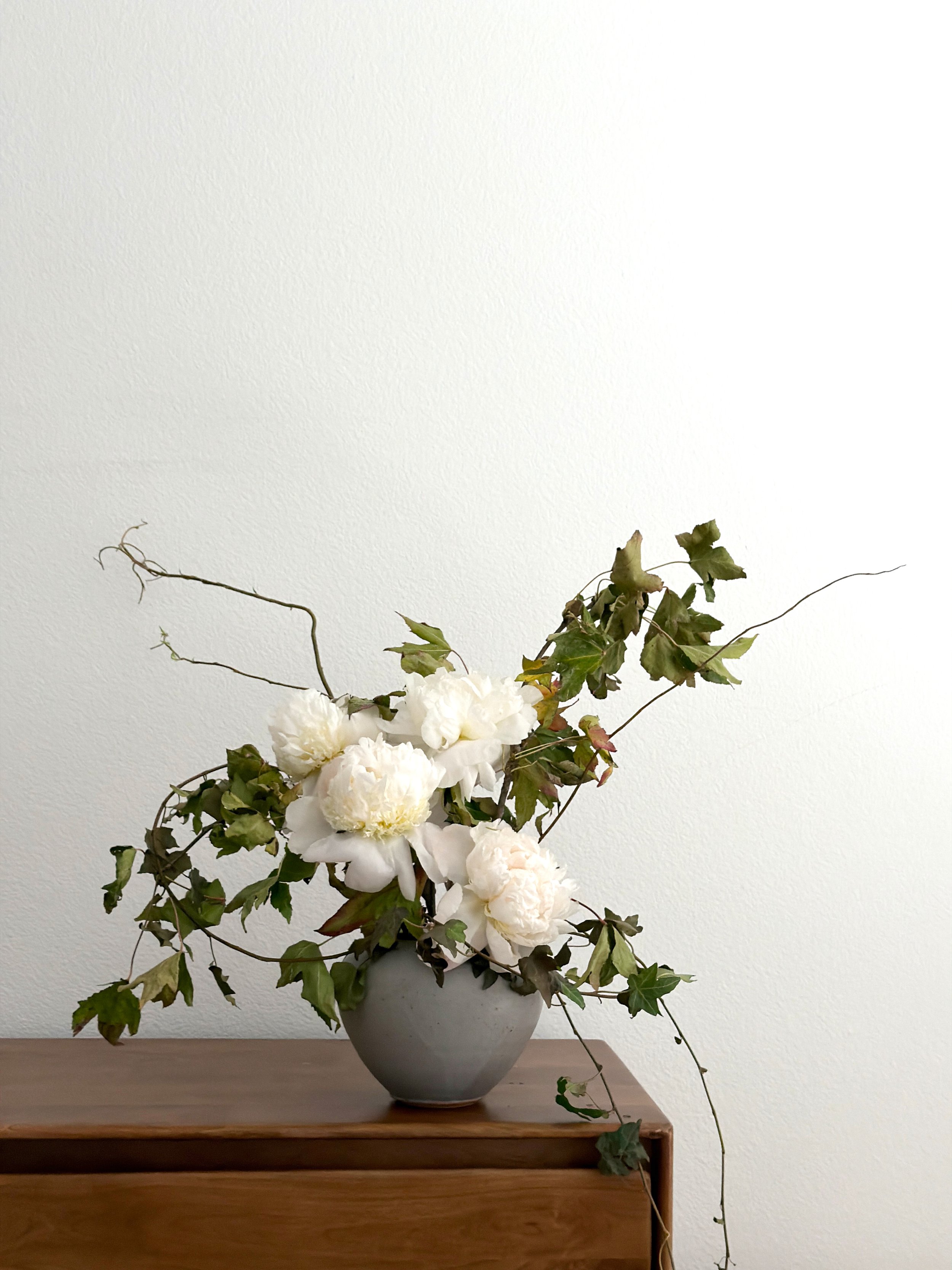 A grey vase with white peony flowers and green ivy leaves on a wooden surface against a plain white wall.