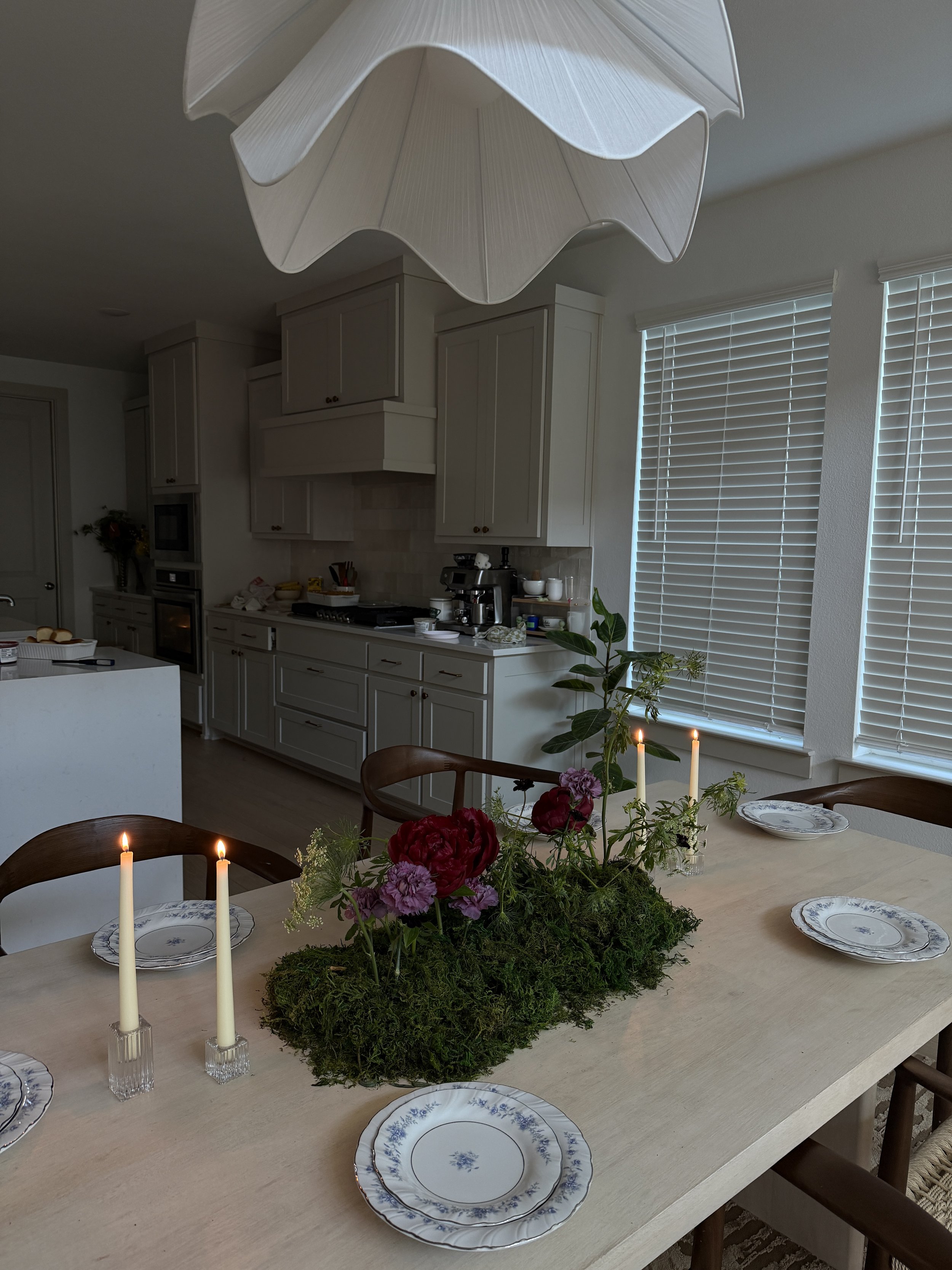 A dining table decorated with a moss and flower centerpiece, surrounded by plates and candles in a bright kitchen with white cabinets and window blinds.