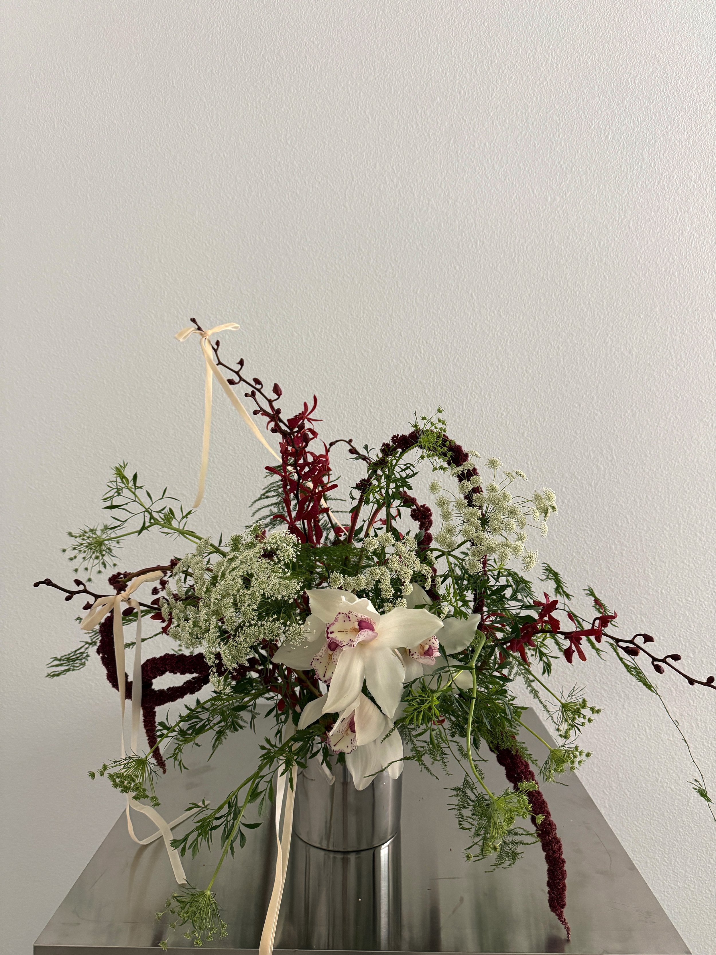 A floral arrangement in a metallic container with white and deep red flowers, including orchids and other greenery, tied with cream-colored ribbons.
