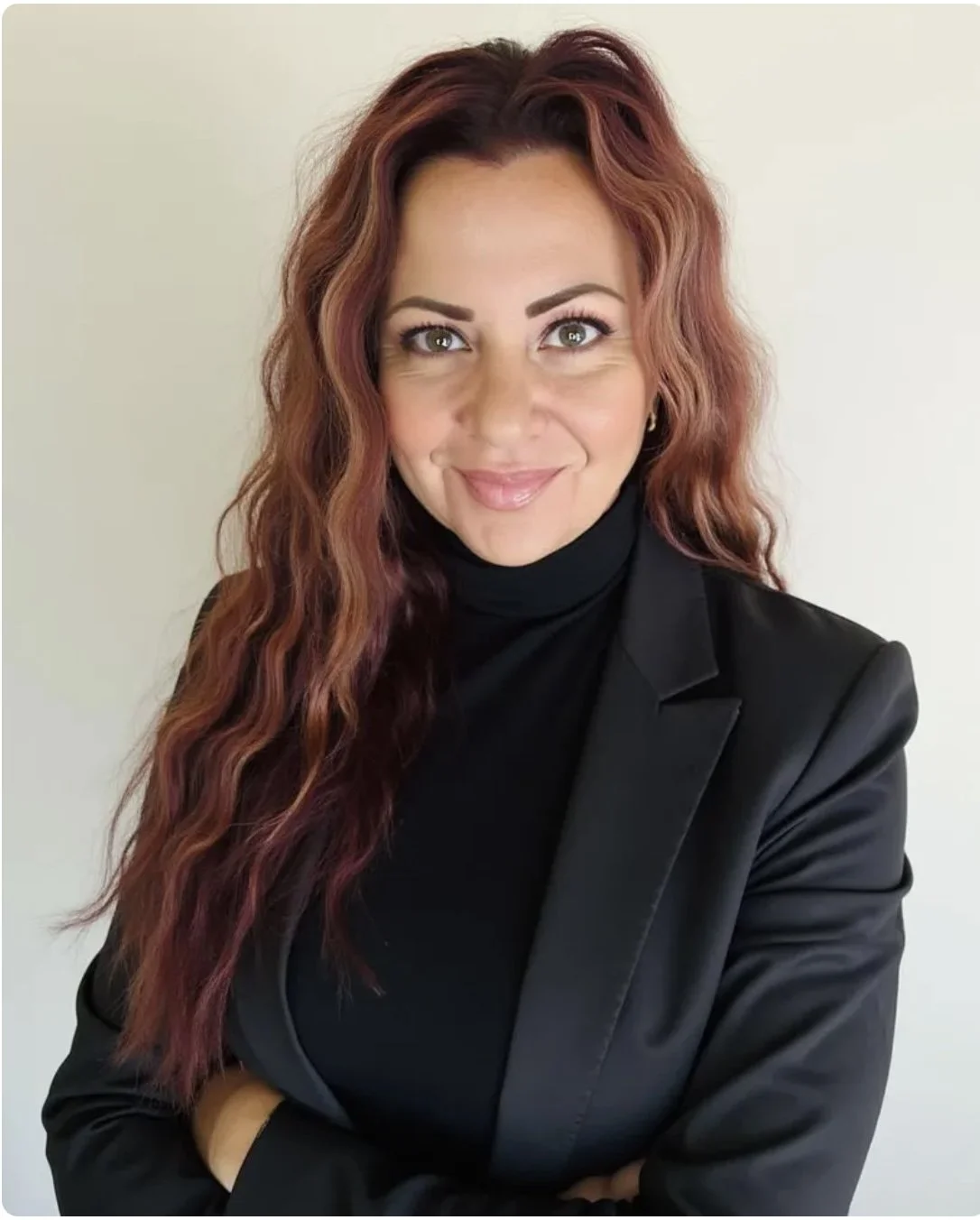 A woman with long wavy burgundy hair, wearing a black blazer and turtleneck, smiling with arms crossed in front of a plain light-colored background.
