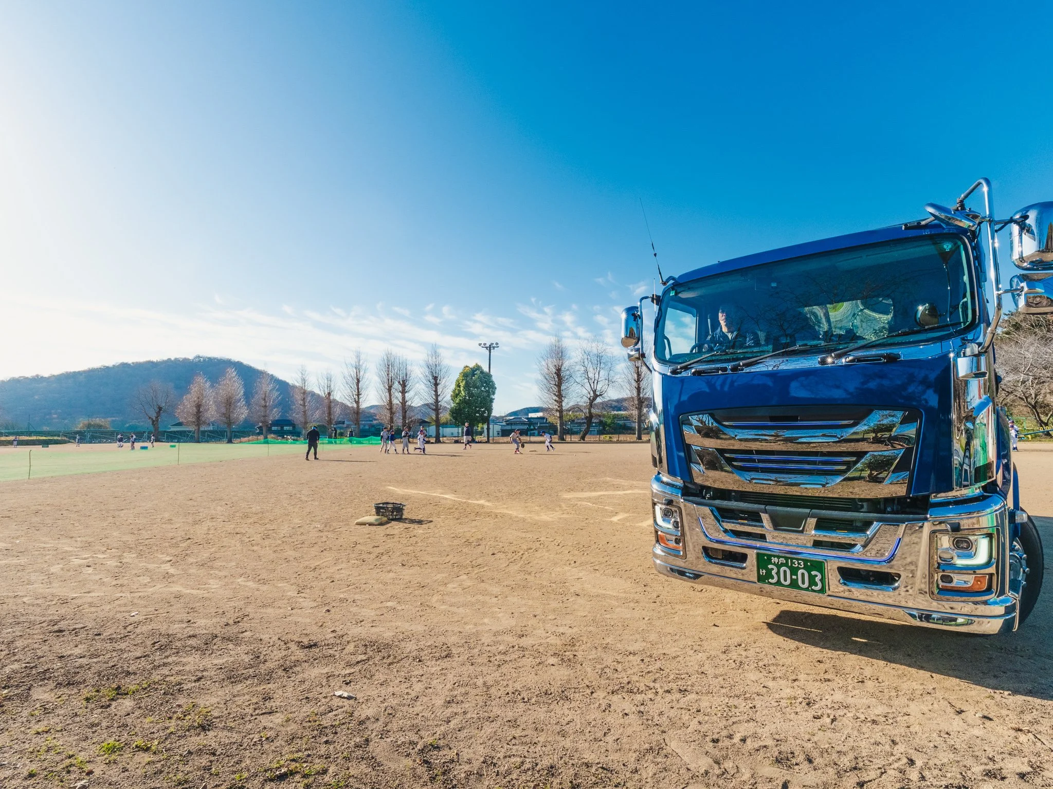 野球場の土のグラウンドと、バスの側面を映した風景。遠くで子供たちが野球をしているのが見える。