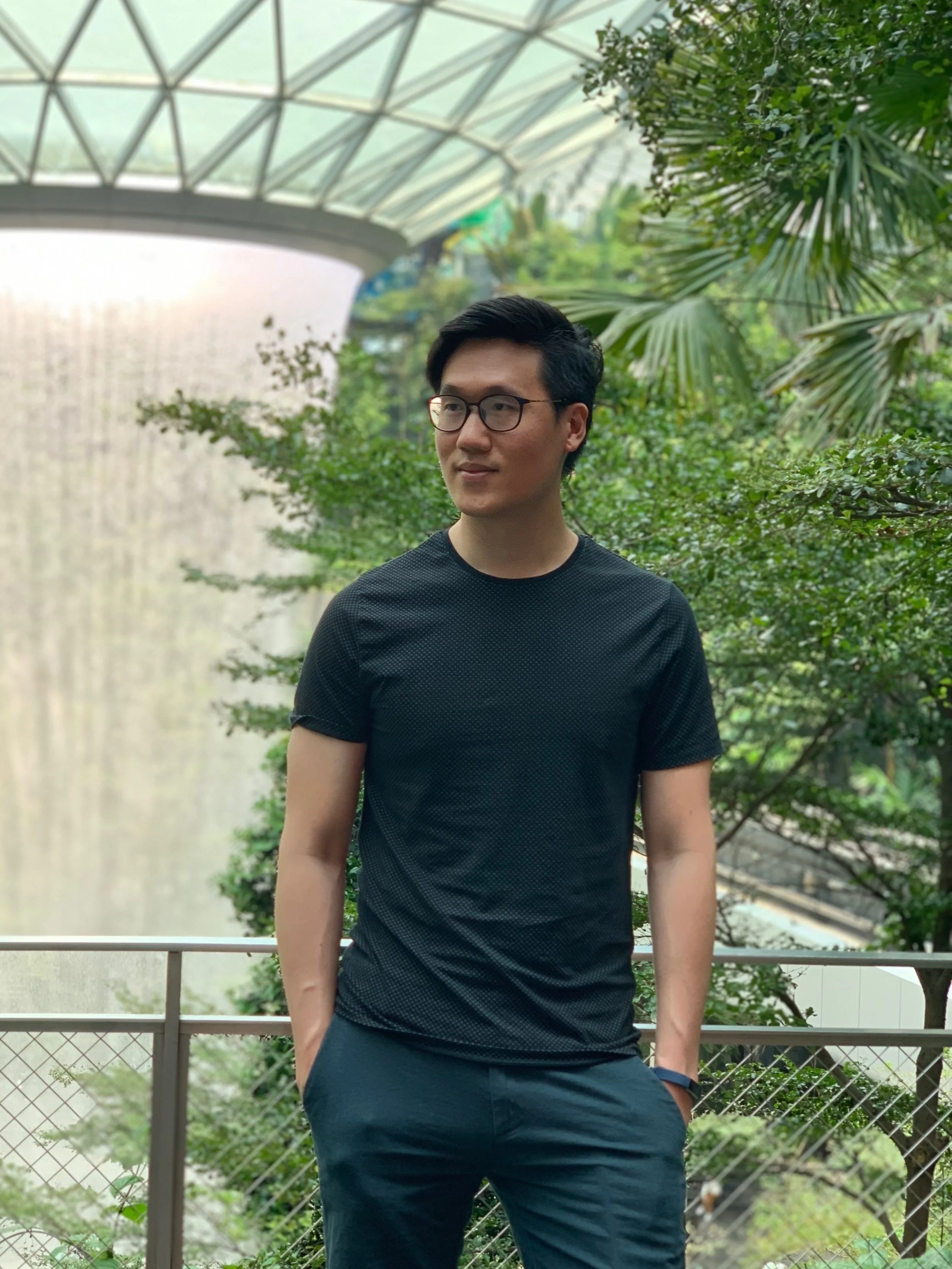 Deacon Geoffrey Goh, a young man with black hair and glasses standing outdoors with lush green plants in the background and a large glass-domed structure overhead.