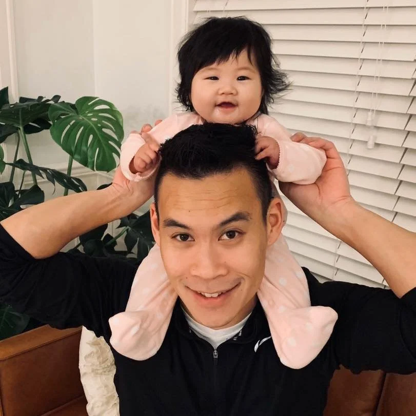 Deacon Erik Lew, A young man with short black hair, smiling and holding a baby girl on his shoulders; she has medium-length black hair, wearing a pink outfit, smiling. They are inside a room with white blinds and a plant with large green leaves.