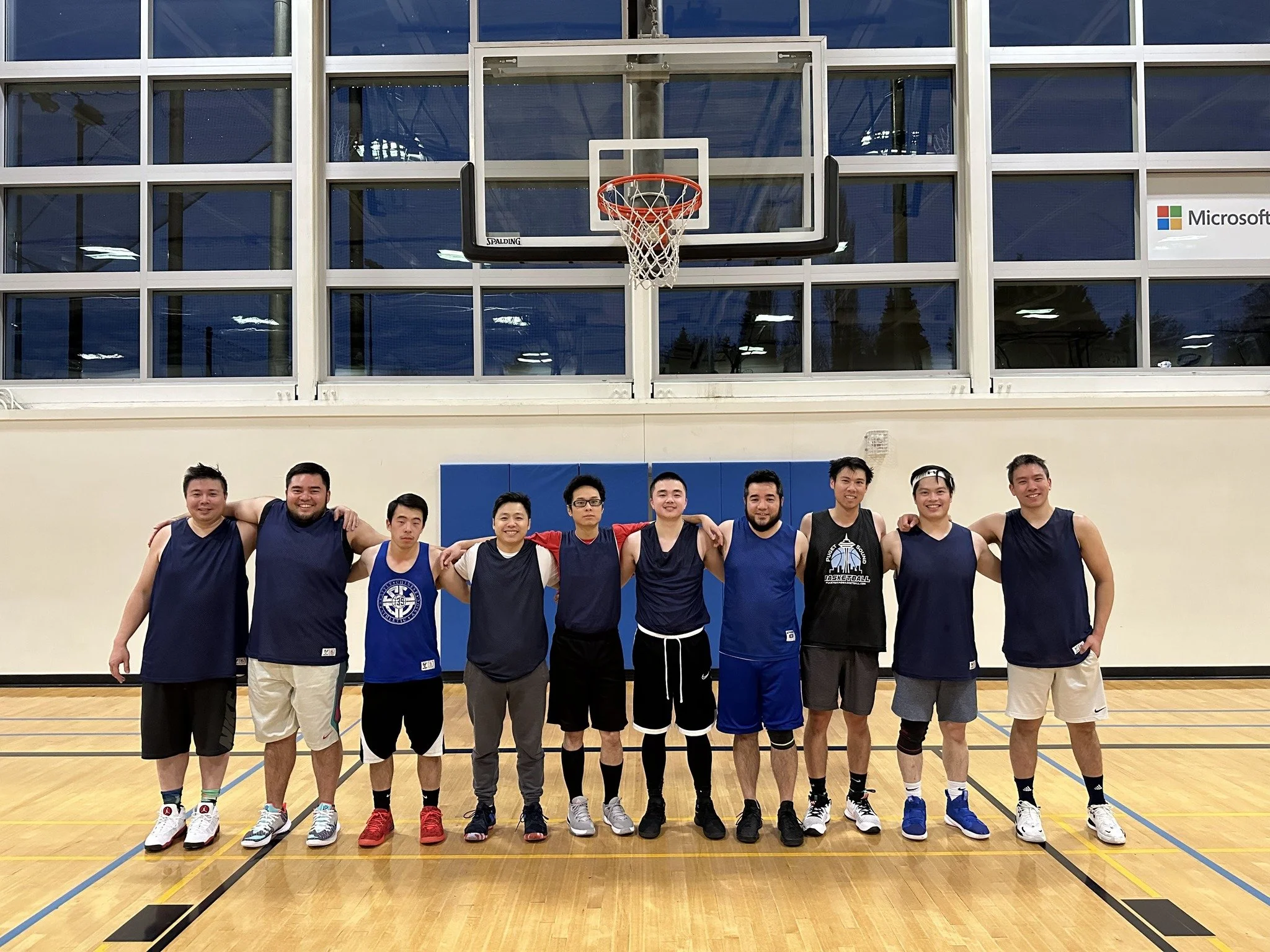 A group of ten young men standing arm-in-arm on a basketball court inside a gymnasium with a basketball hoop overhead, posing for a team photo.