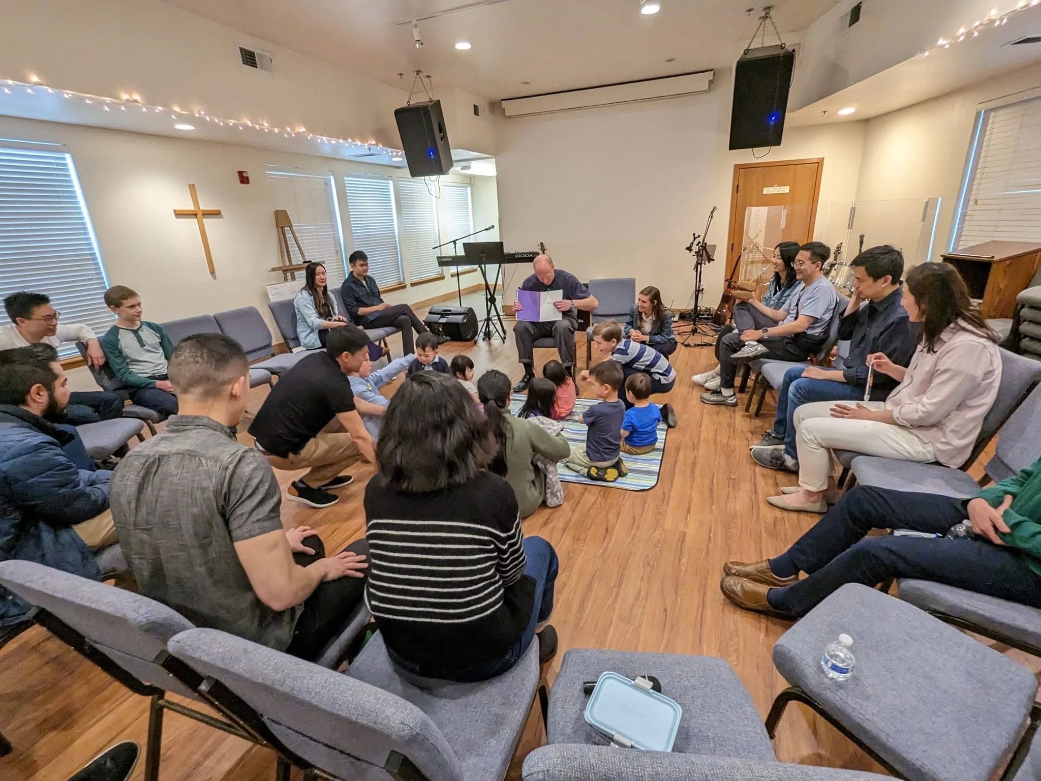 A group of children and adults sitting on the floor and chairs in a church or community room, listening to a bald man reading from a book. There are musical instruments, speakers, and a cross on the wall.