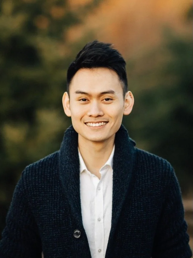 A smiling young man with short black hair wearing a white shirt and dark knit cardigan outdoors with blurred trees in the background.