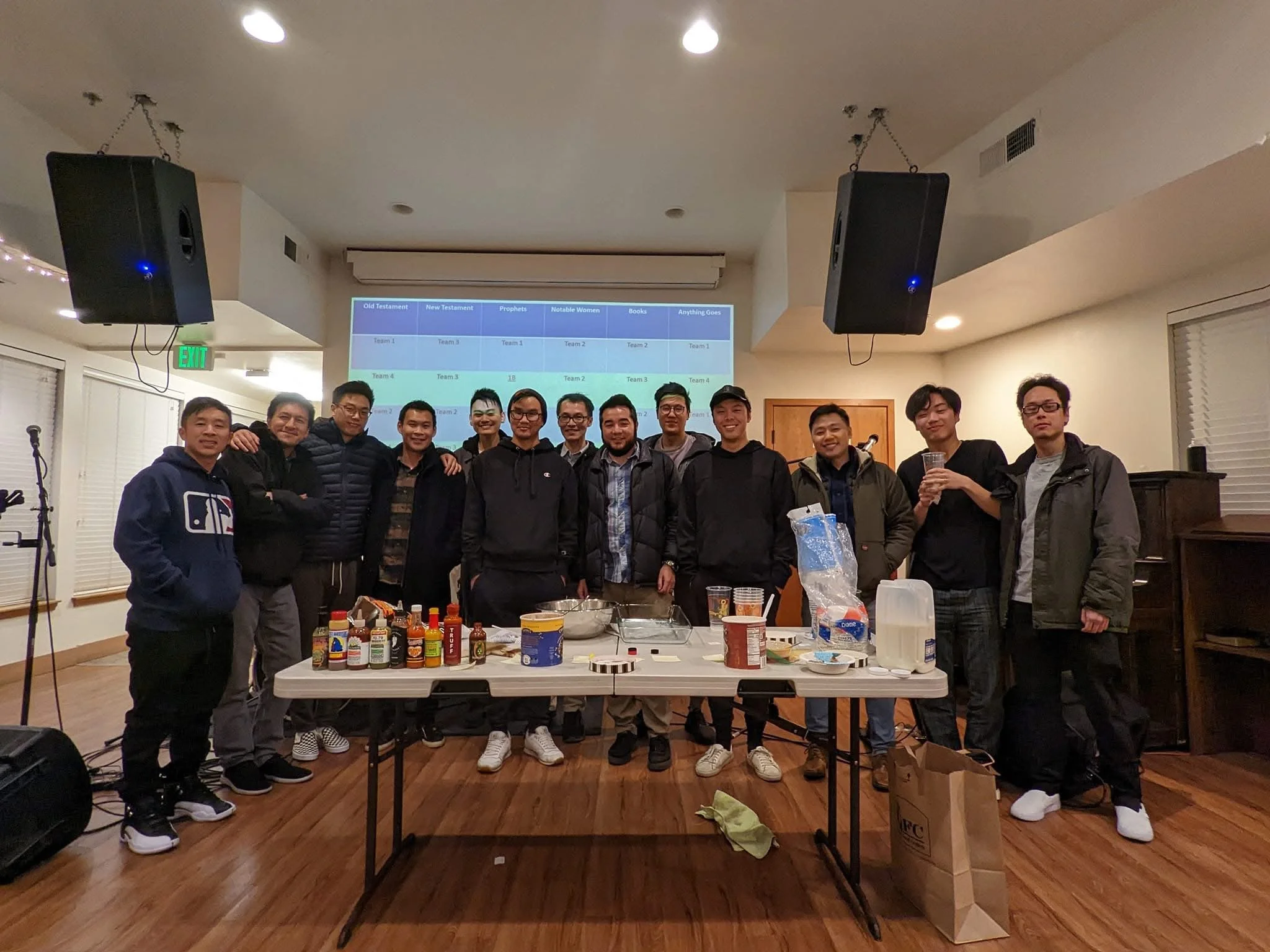 Group of 14 people standing behind a table with bottles of hot sauce and food, posing indoors in a room with wood flooring and speakers, with a large projected chart on the wall behind them.