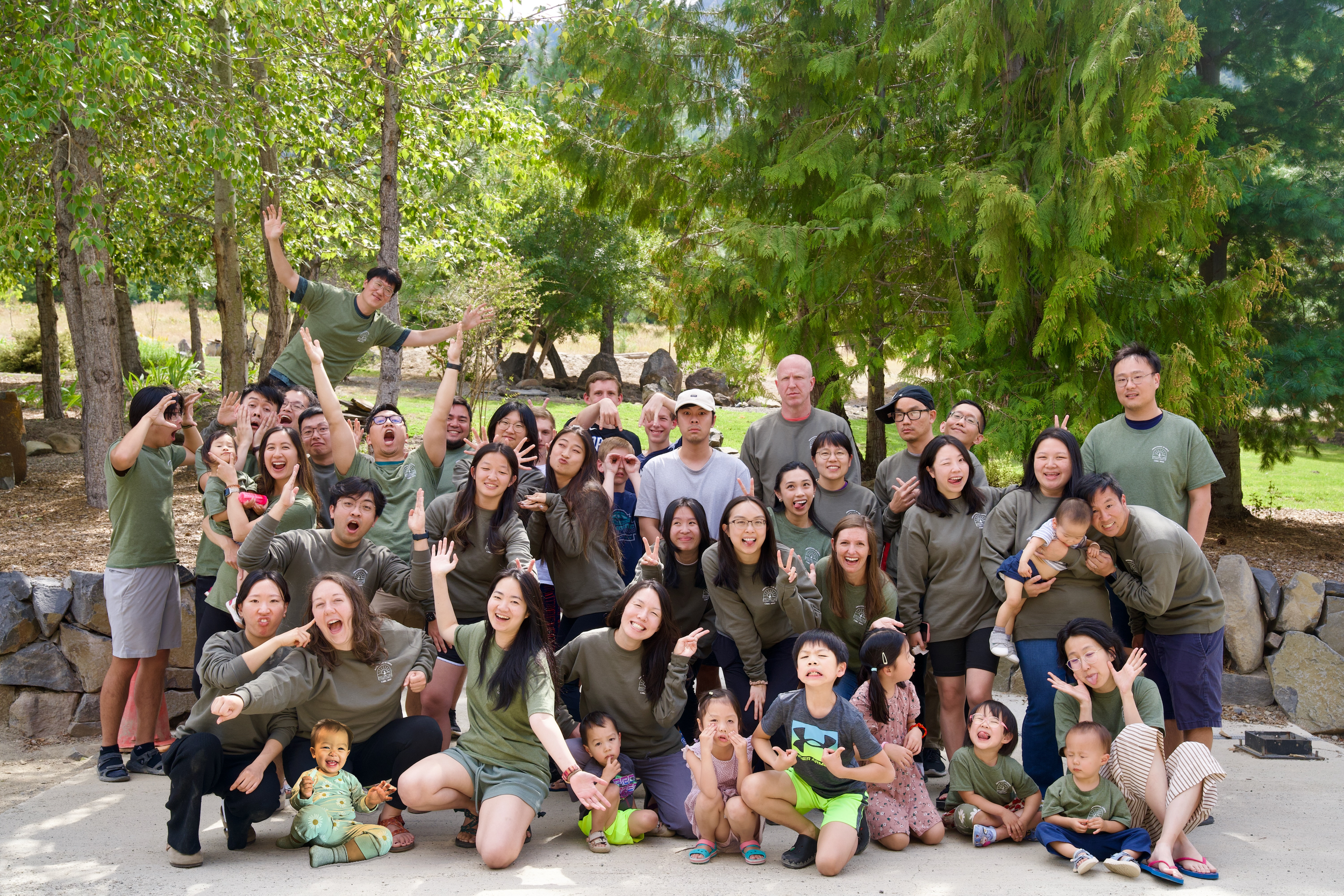 A large group of children and adults outdoors posing for a photo in front of trees, with some making peace signs, smiling, and several children sitting or standing in front.