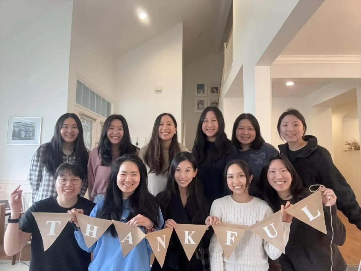 A group of twelve women smiling and holding a brown banner spelling 'THANKFUL' in a living room with white walls and framed pictures.