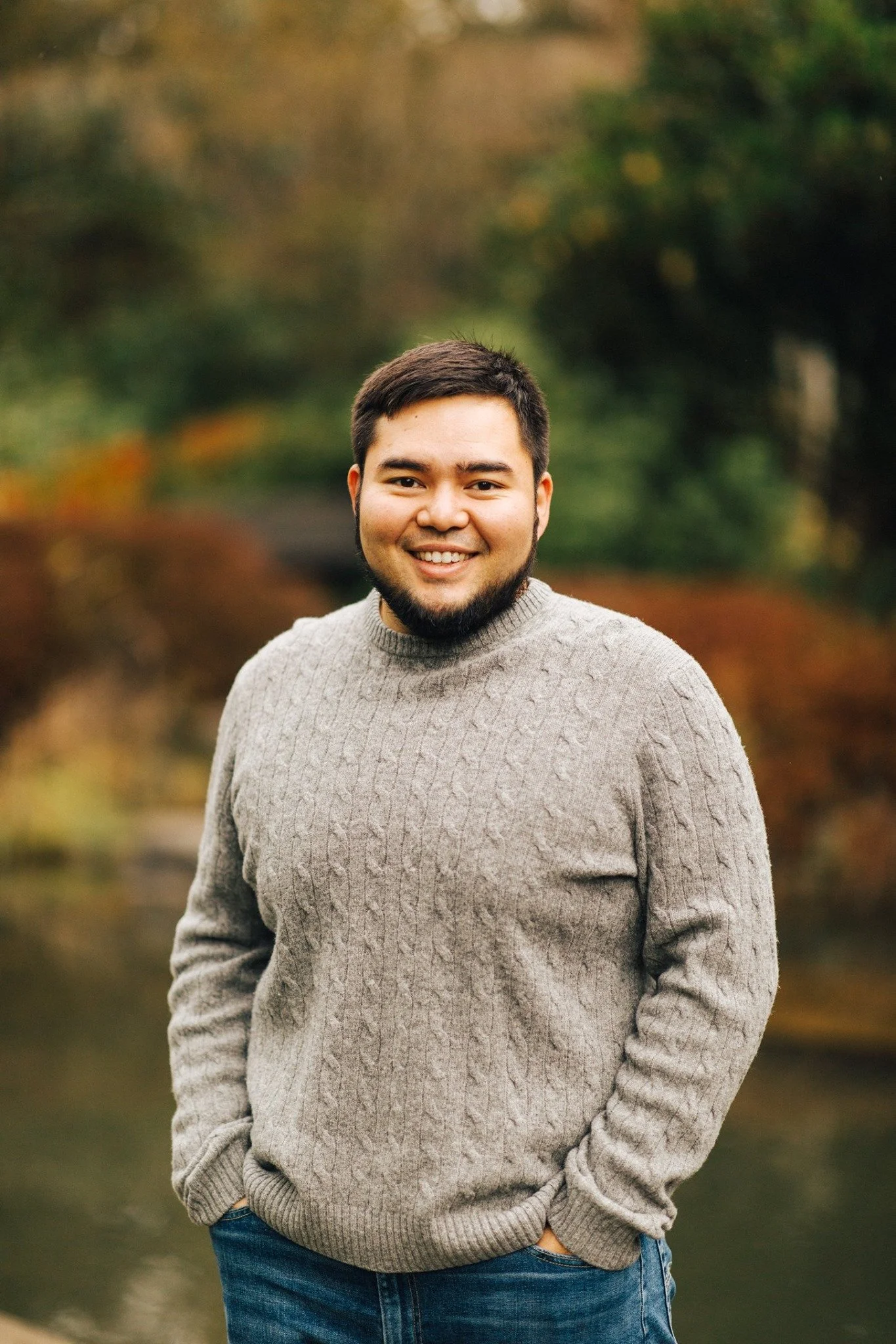 Deacon Nicholas Pleasants, a young man with dark hair and beard smiling outdoors in a park during autumn, wearing a gray cable-knit sweater and blue jeans.