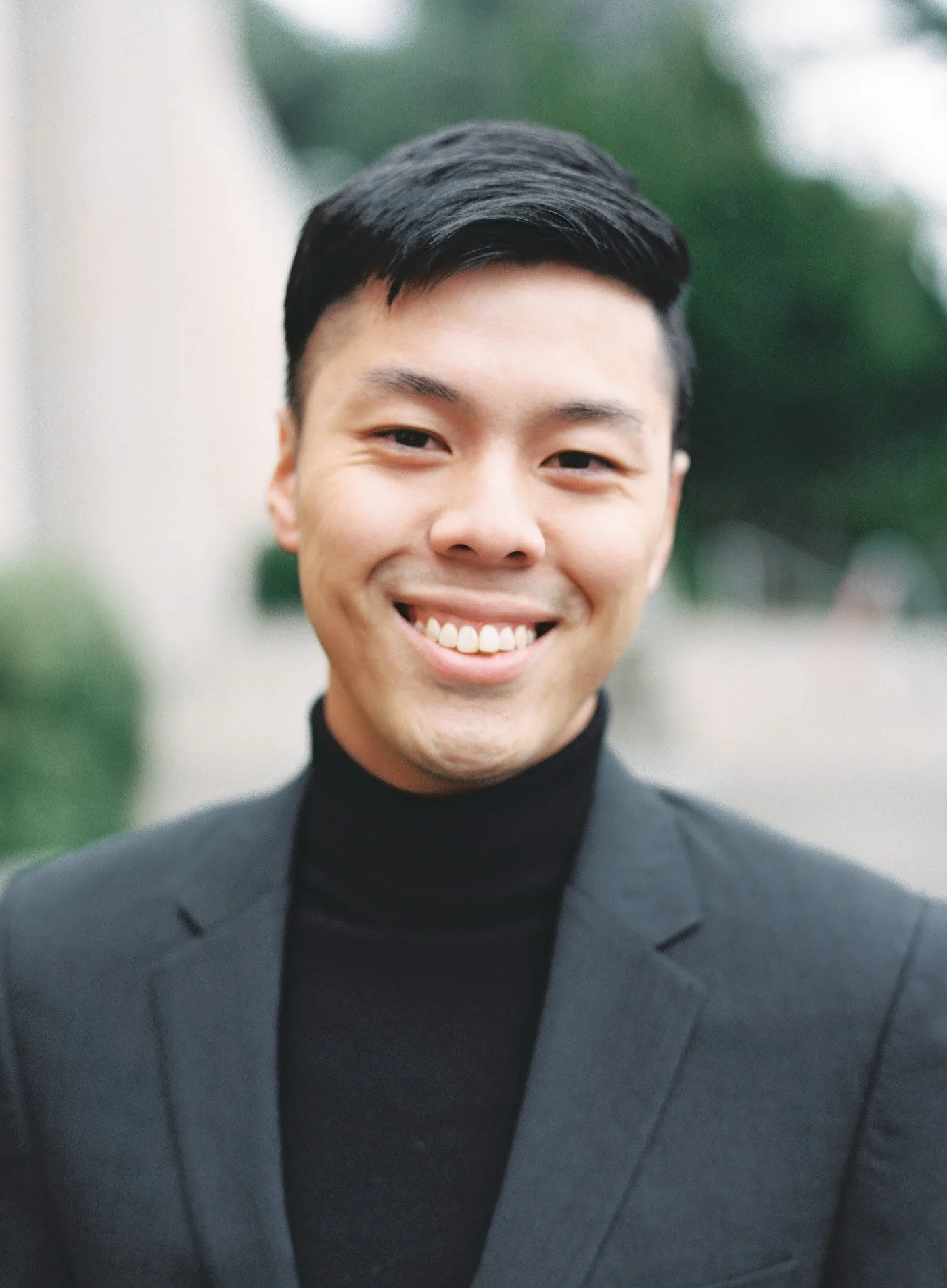 A smiling young Asian man in a black turtleneck and gray blazer outdoors.