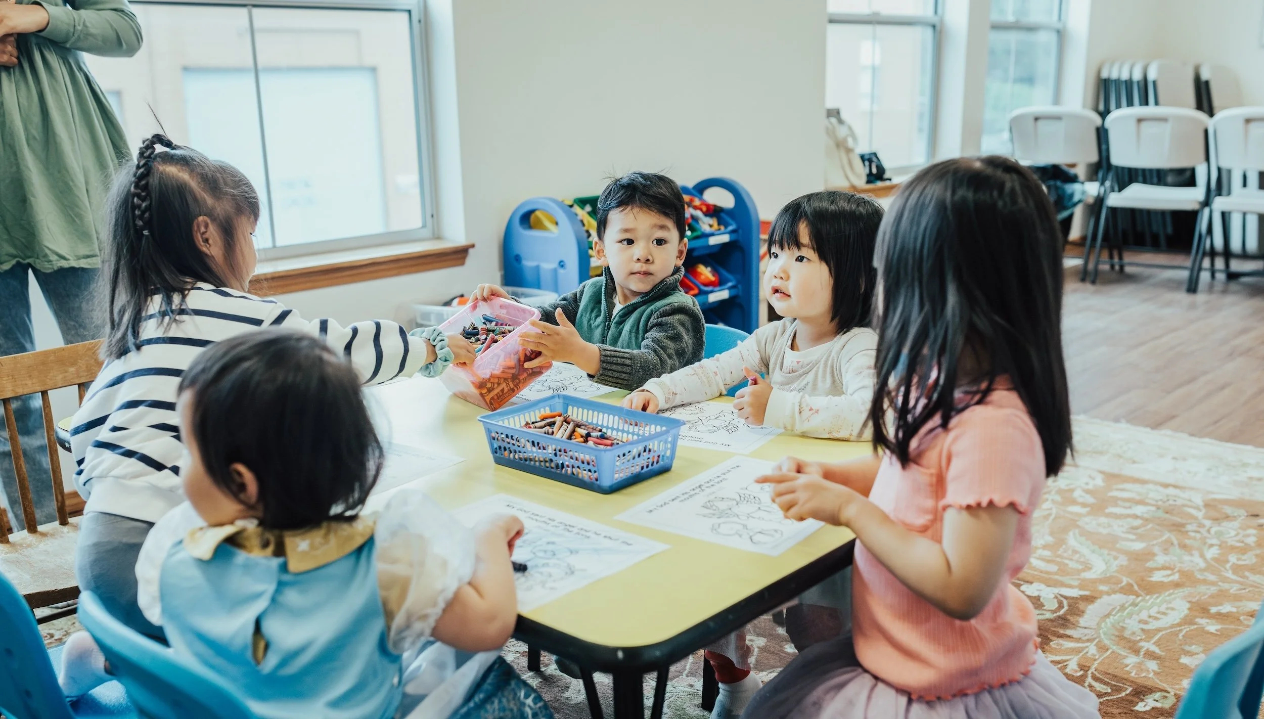 Group of young children sitting around a yellow table in a classroom, coloring and engaging with each other.