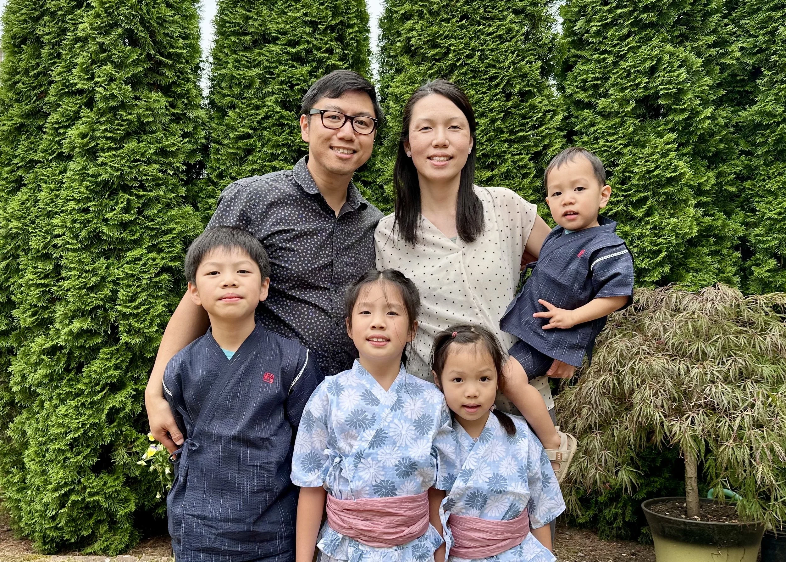 A family of six posing outdoors in front of greenery, with two adults and four children dressed in traditional Japanese yukatas and casual clothing.