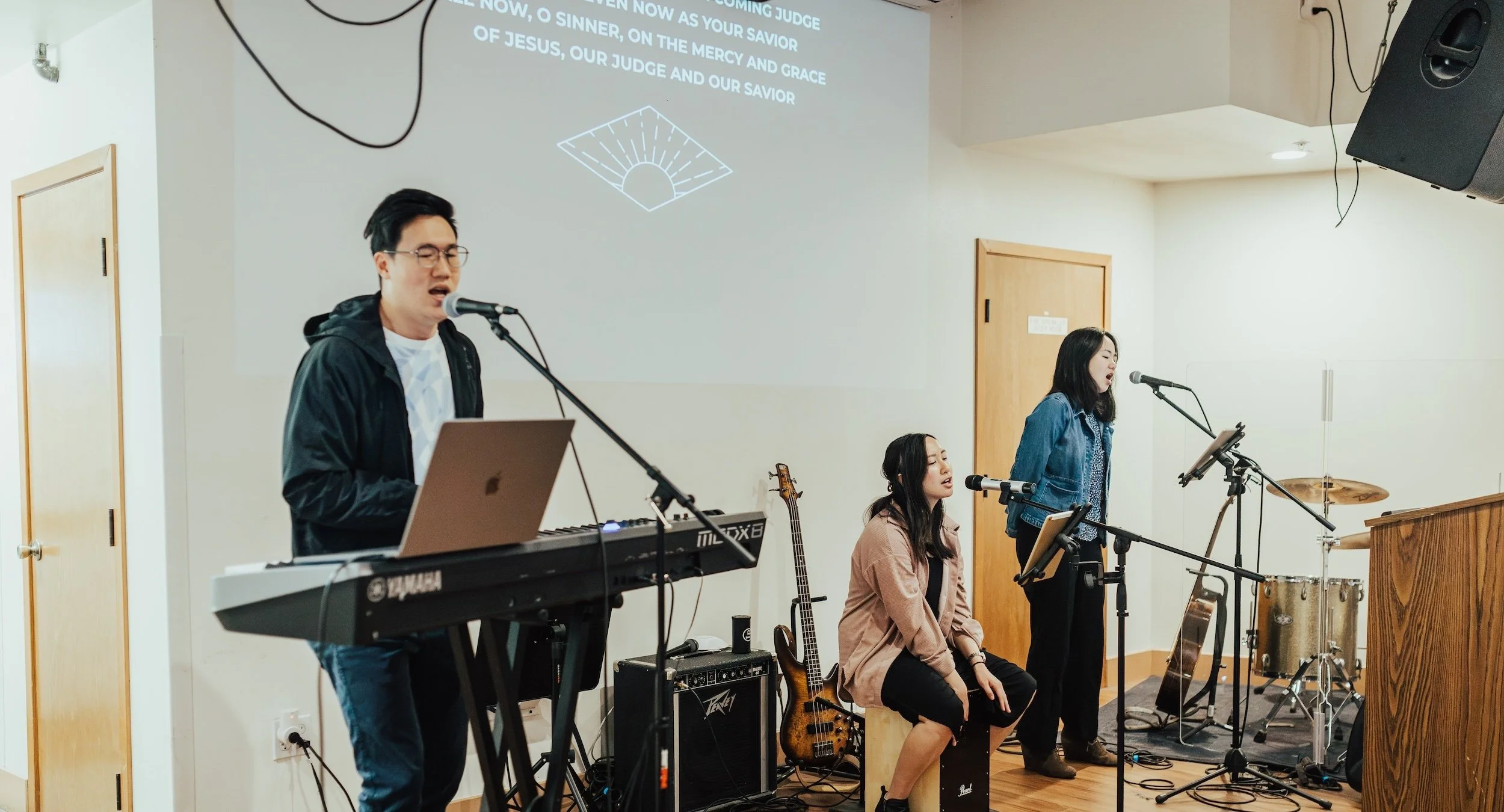 Three young people singing and playing music in a church or practice room, with a keyboard, microphone, guitar, and drums.