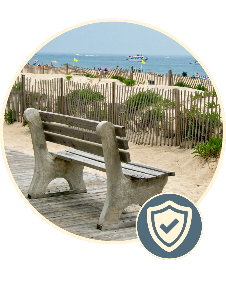 Empty wooden bench on the beach with sand, green plants, wooden fences, and ocean with boats and a clear sky in the background.