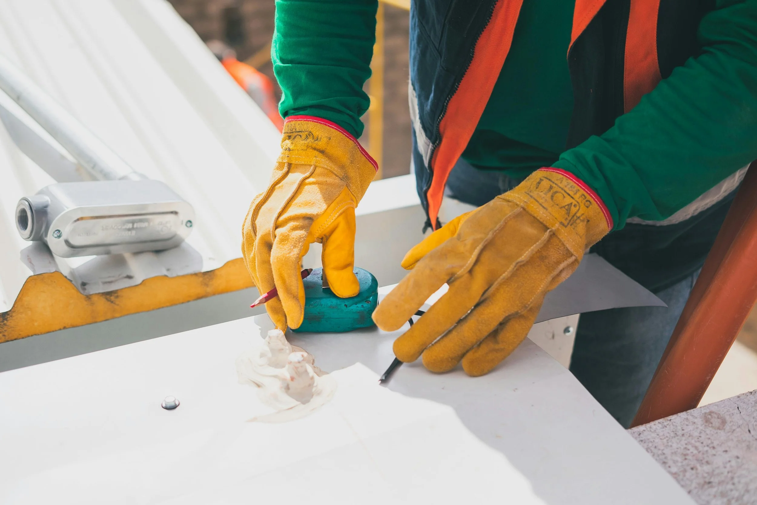 Construction worker wearing yellow gloves and a green shirt, marking a white surface with a black marker, with a level tool and a sealed tube nearby.
