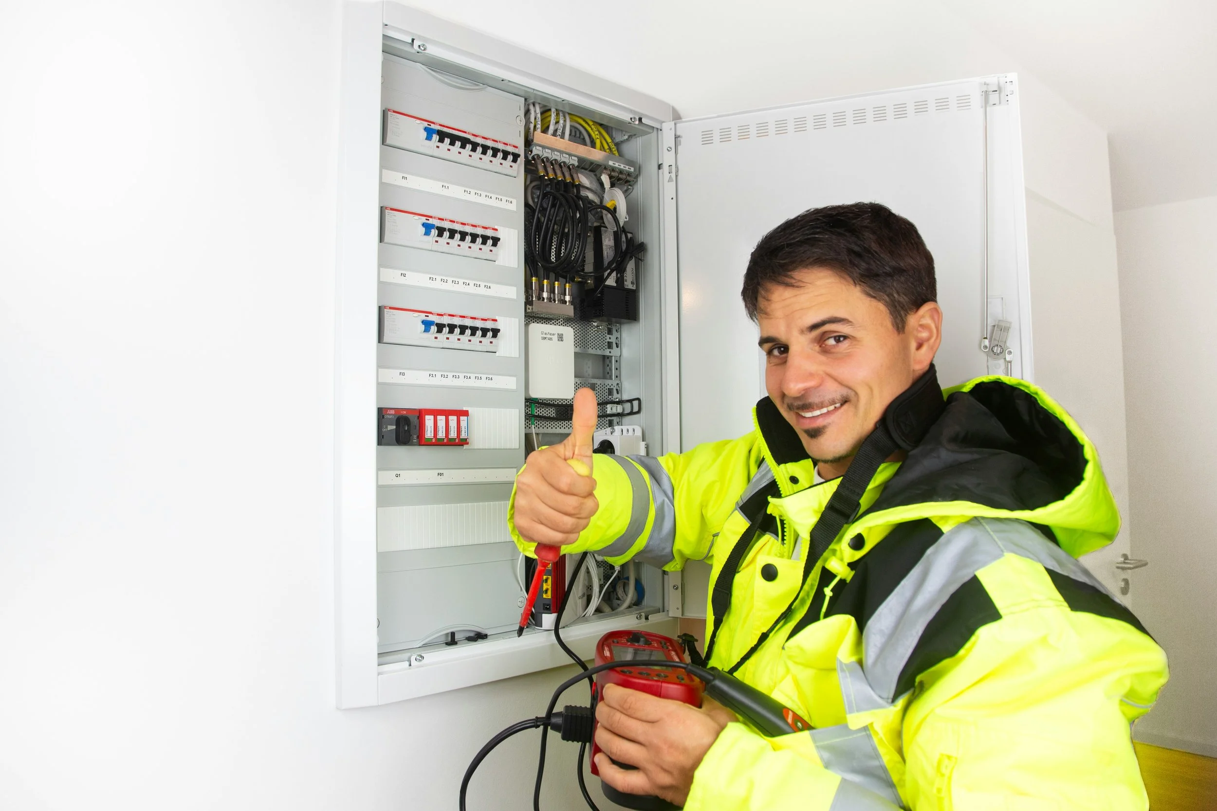 Electrician inspecting an electrical panel with a multimeter, giving a thumbs up and smiling.