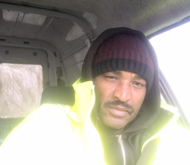 A man wearing a black beanie and a yellow jacket sits inside a vehicle, looking slightly to the side.